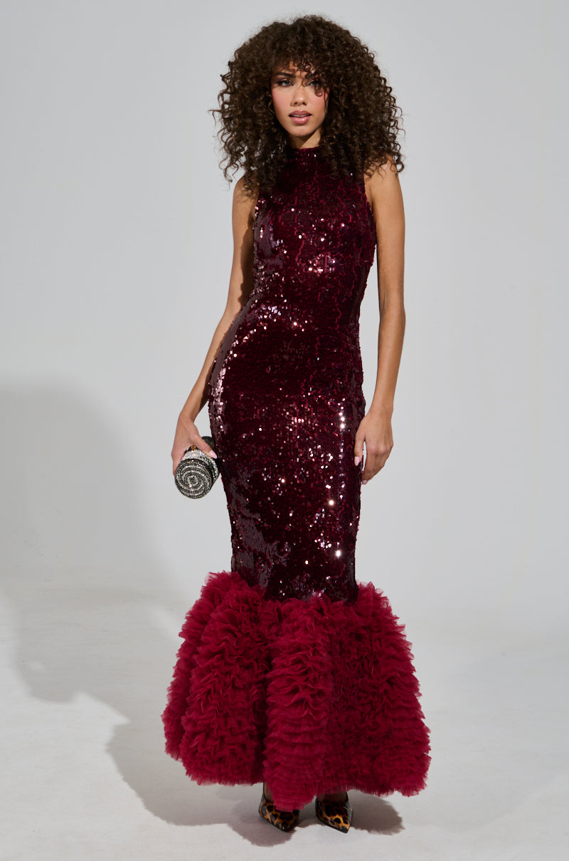 A woman wearing a shimmering burgundy Belle Of The Ball Sequin Ruffle Maxi Dress, characterized by an ornate ruffled hem, holds a small silver clutch. With curly hair and a confident pose, she stands against a plain background in this elegant mermaid silhouette.