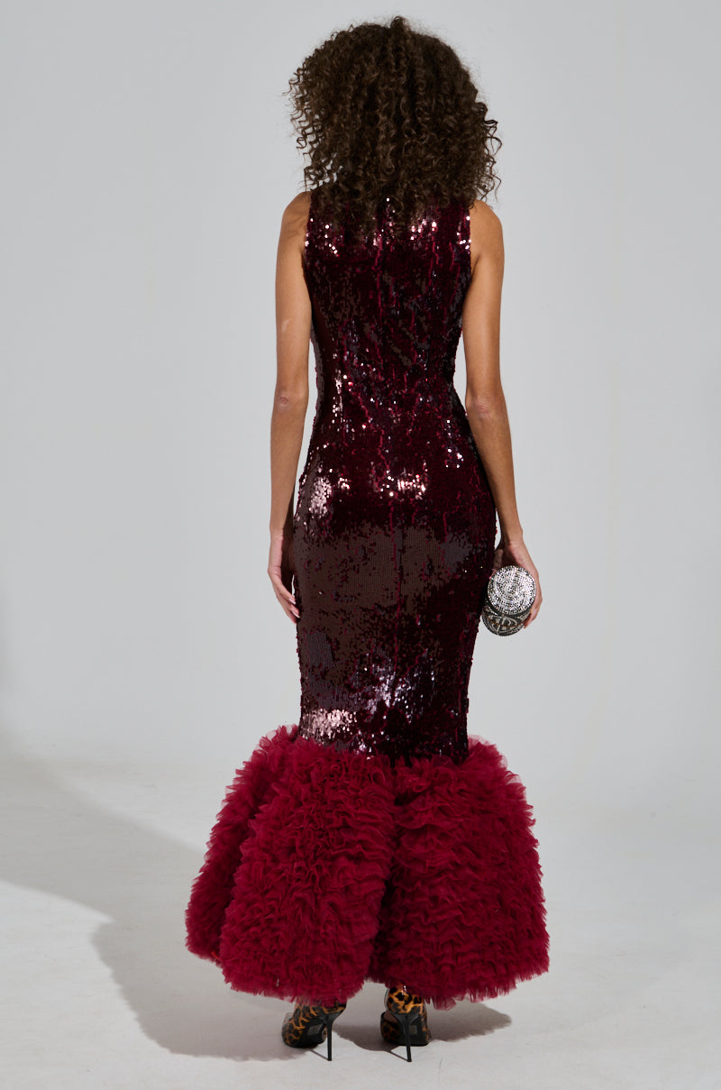A woman with curly hair is wearing the shimmering burgundy Belle of the Ball Sequin Ruffle Maxi Dress, featuring a dramatic feathered hem. She holds a small round clutch in her right hand and stands against a plain background, facing away from the camera.