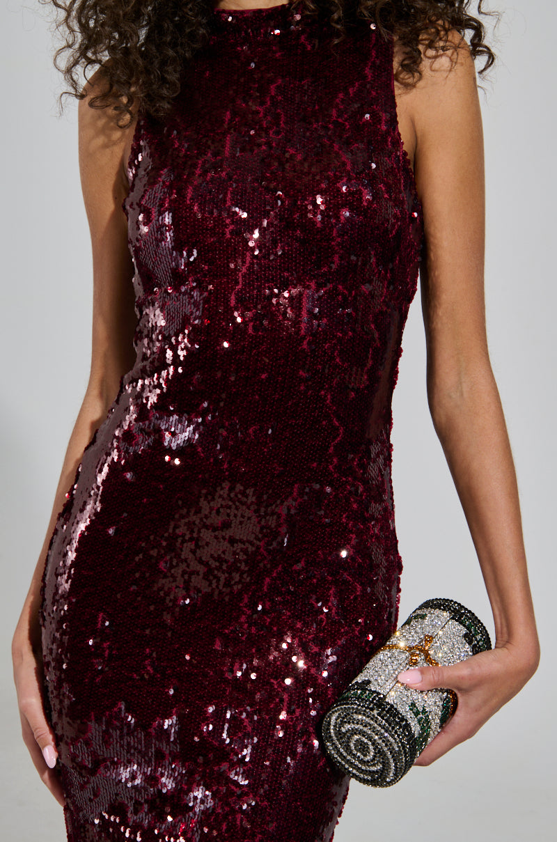 Dressed in a sleeveless, BELLE OF THE BALL SEQUIN RUFFLE MAXI DRESS, a person holds a small, patterned clutch. The dark red, form-fitting gown glimmers under the light like a dazzling mermaid silhouette. The clutch showcases an intricate design with contrasting colors.