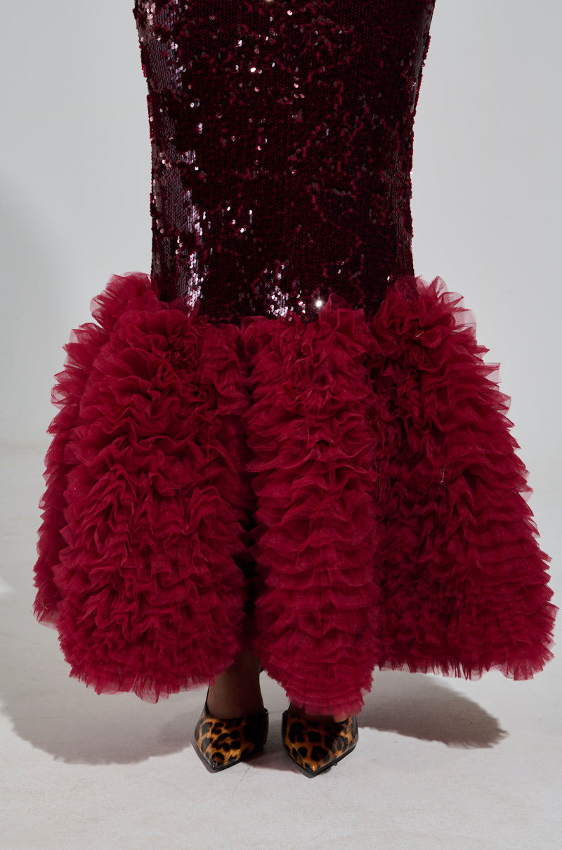 A person wearing a burgundy Belle Of The Ball Sequin Ruffle Maxi Dress with a voluminous ruffled hem stands on a light background. The dress, featuring a mermaid silhouette, is paired with pointed leopard print shoes.