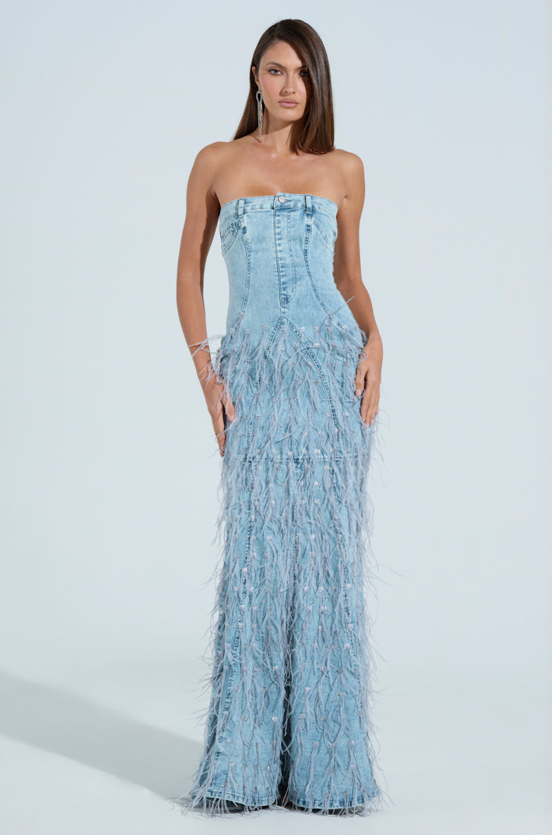 A woman poses in a BRITTANY FEATHER DENIM MAXI DRESS by AKIRA Label, showcasing its strapless design with frayed feather-like textures from the waist down. The dress's straight silhouette flatters her figure, complemented by her long hair and hoop earrings against a plain white background.