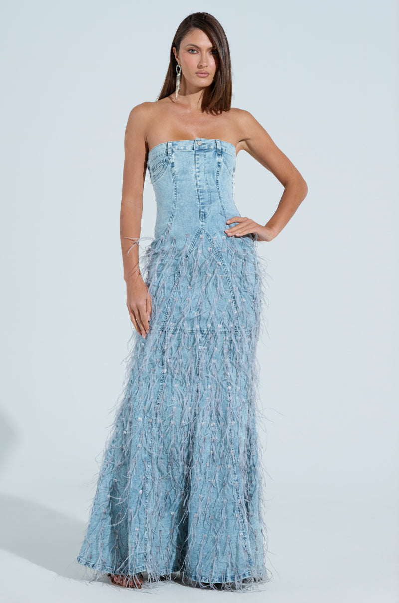 A woman poses in a strapless BRITTANY FEATHER DENIM MAXI DRESS against a light backdrop. Her straight hair falls to one side, complemented by earrings. The dress features a fitted bodice and textured, feather-like skirt, perfectly blending chic style with elegance.