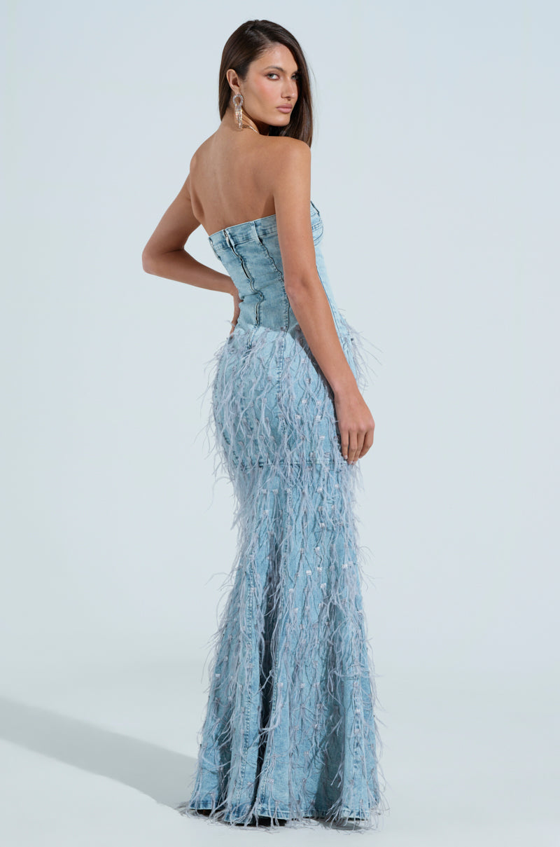 A woman in a strapless BRITTANY FEATHER DENIM MAXI DRESS stands against a plain background. Her long hair drapes over one shoulder, and she wears drop earrings. She faces the camera with a confident pose.