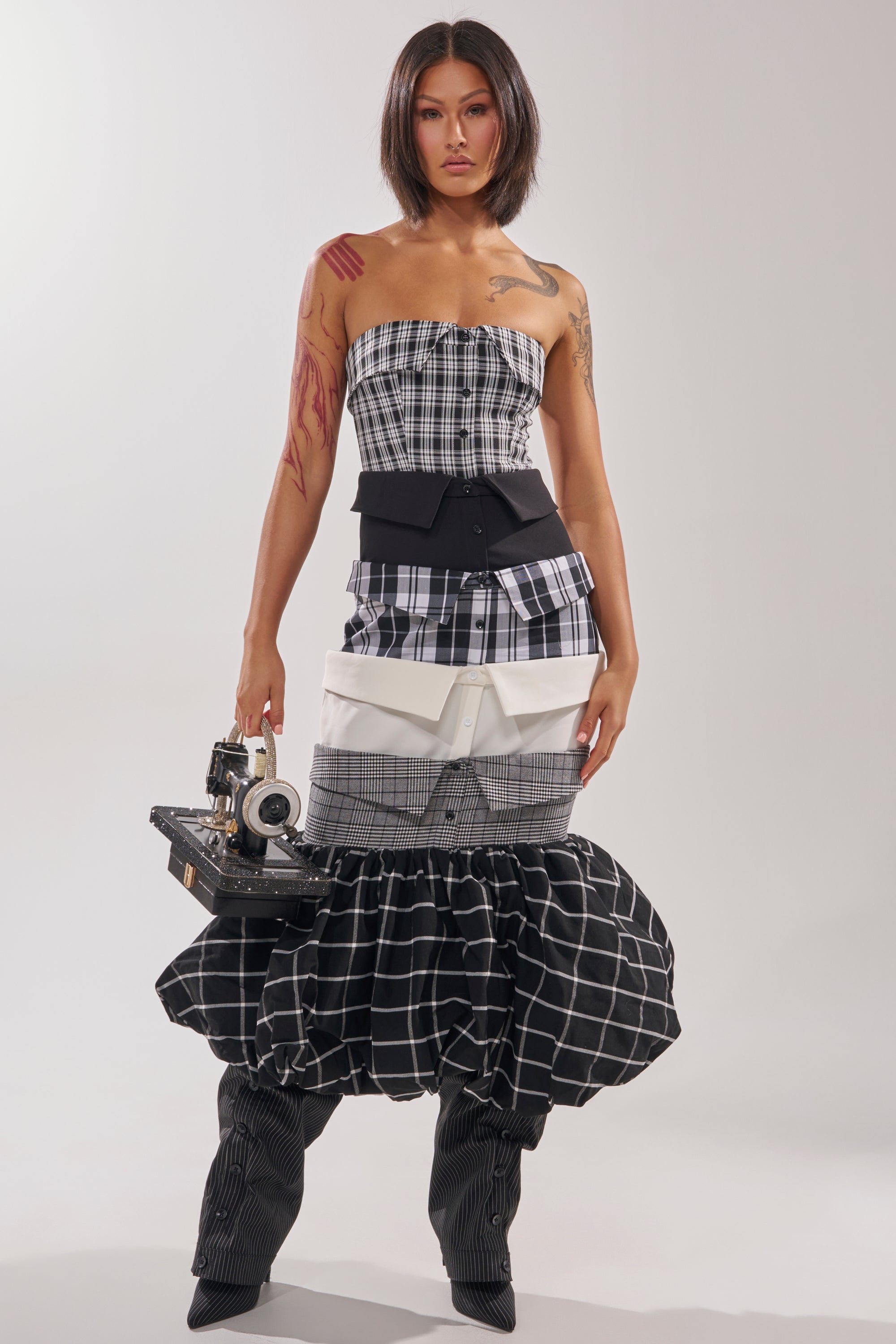 A woman with a bob hairstyle wears the CHANGING IT UP PLAID MIDI DRESS with a strapless, tiered design and matching plaid boots. She holds a vintage camera, revealing arm tattoos.