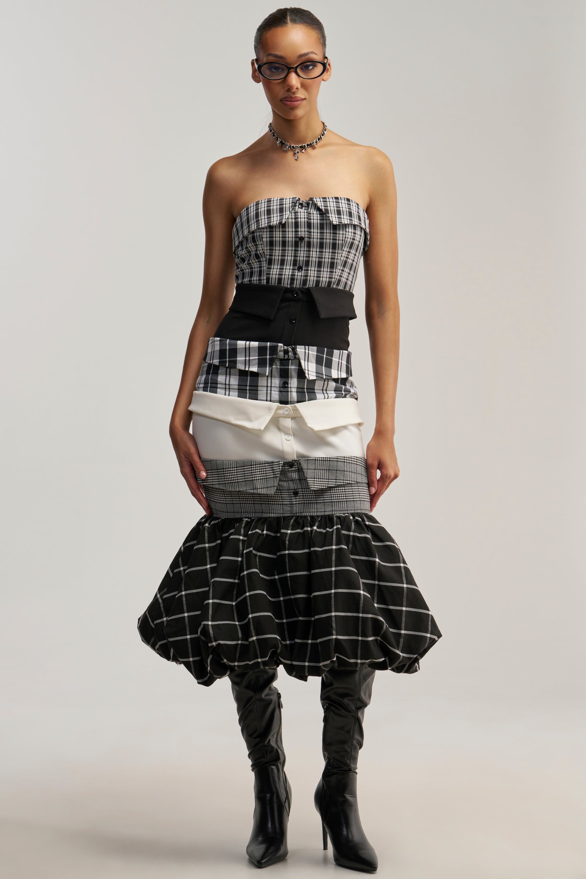 A woman wearing glasses stands against a plain background, dressed in the CHANGING IT UP PLAID MIDI DRESS. This strapless midi dress features a bubble hemline, while its layered skirt—crafted from an assortment of black, white, and plaid fabrics—adds visual interest to her look. Black knee-high boots complete her stylish ensemble.