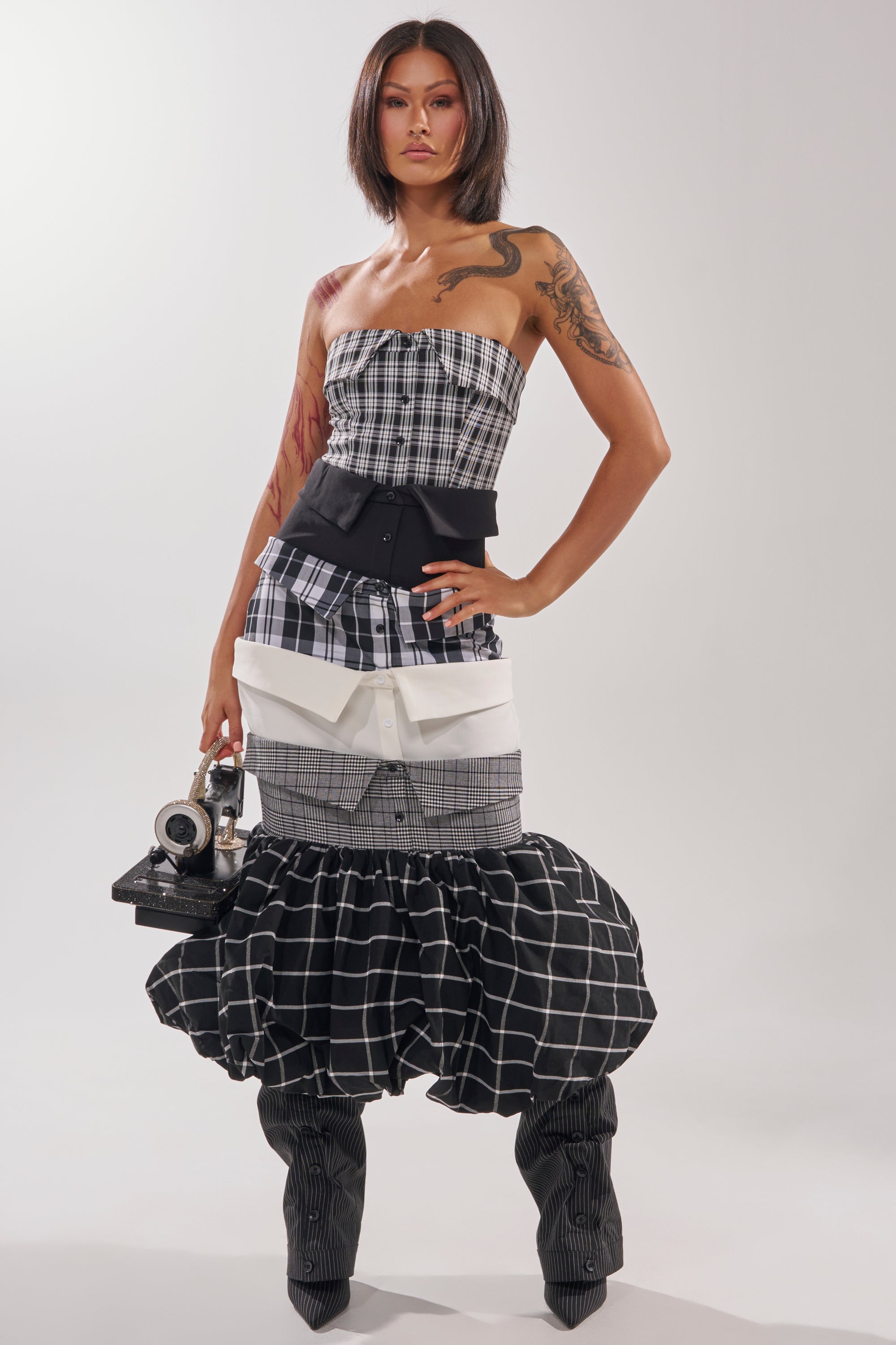 A woman wears the CHANGING IT UP PLAID MIDI DRESS with a strapless neckline, structured black waist corset, large white pockets, and tall black ruched boots while holding a vintage camera against a plain white background.