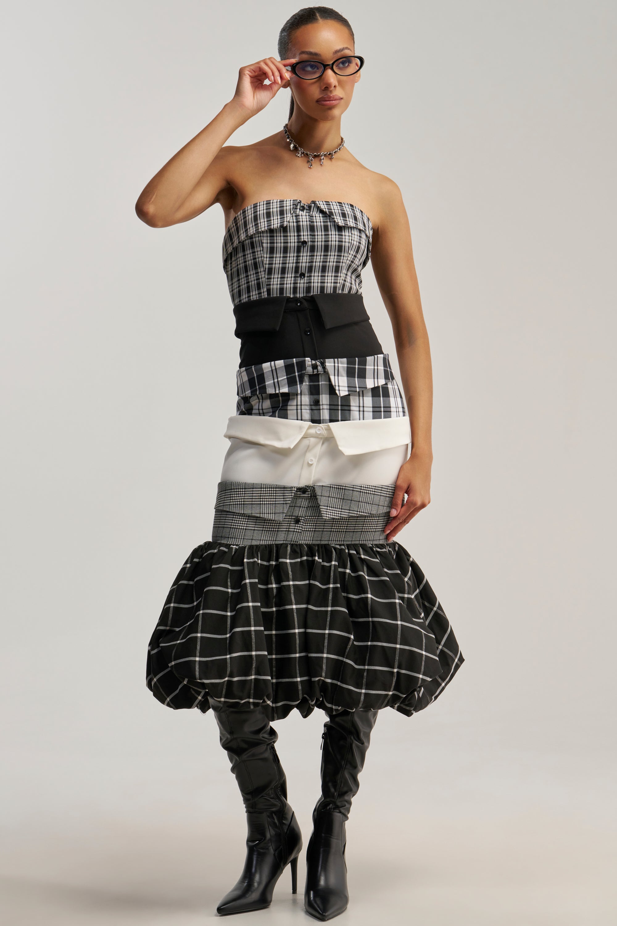Wearing the CHANGING IT UP PLAID MIDI DRESS layered over a shirt, a woman in glasses styles her look with a wide black corset and a tiered checkered skirt featuring a subtle bubble hemline, finishing off with tall black boots as she poses confidently against a plain background.