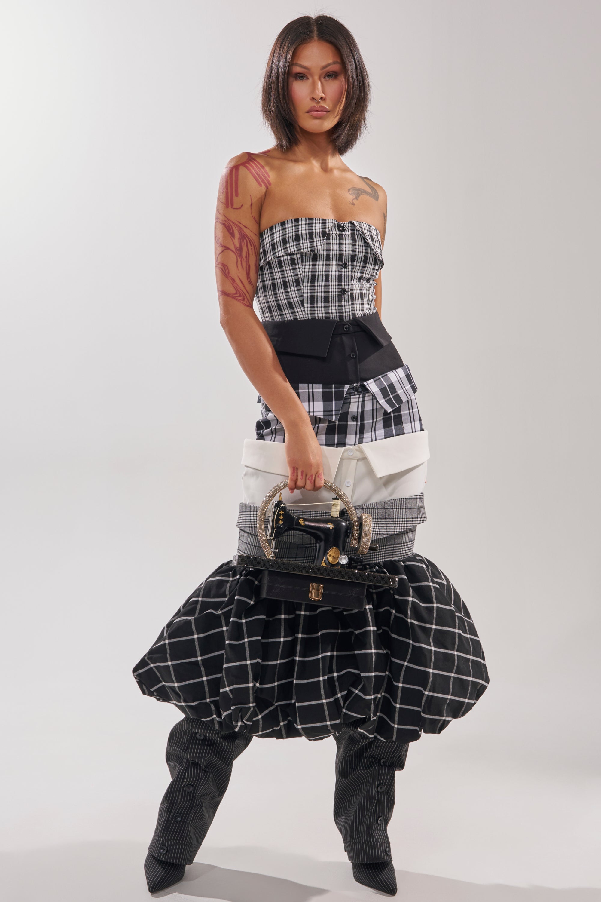 A woman with a bob haircut and arm tattoo stands against a plain background, wearing the CHANGING IT UP PLAID MIDI DRESS with black and white plaid patterns, a black belt, matching slouchy boots, and carrying a coordinating handbag.