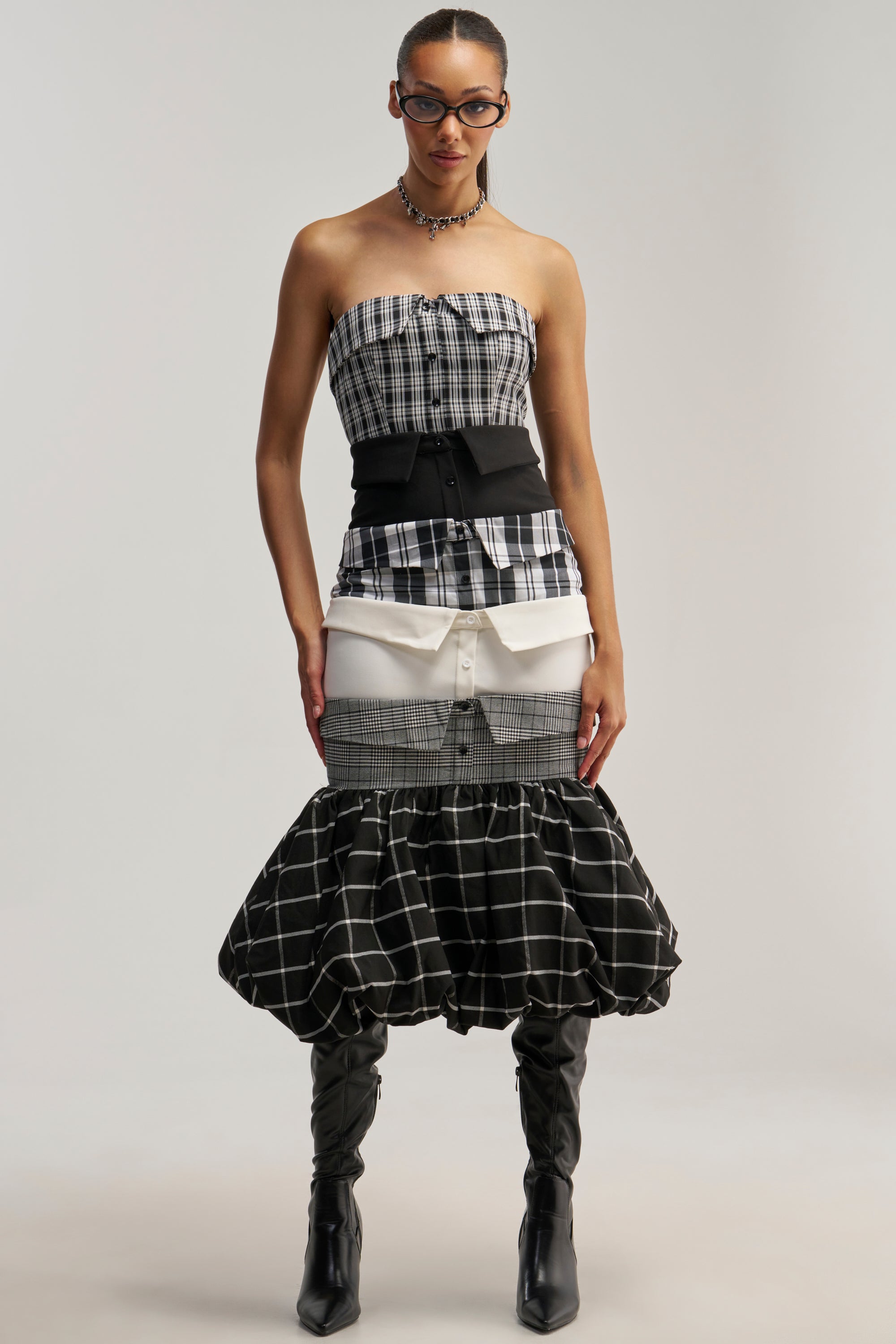 A person wearing glasses and tall black boots stands against a plain background in the CHANGING IT UP PLAID MIDI DRESS, which features a strapless, fitted top and a layered skirt.