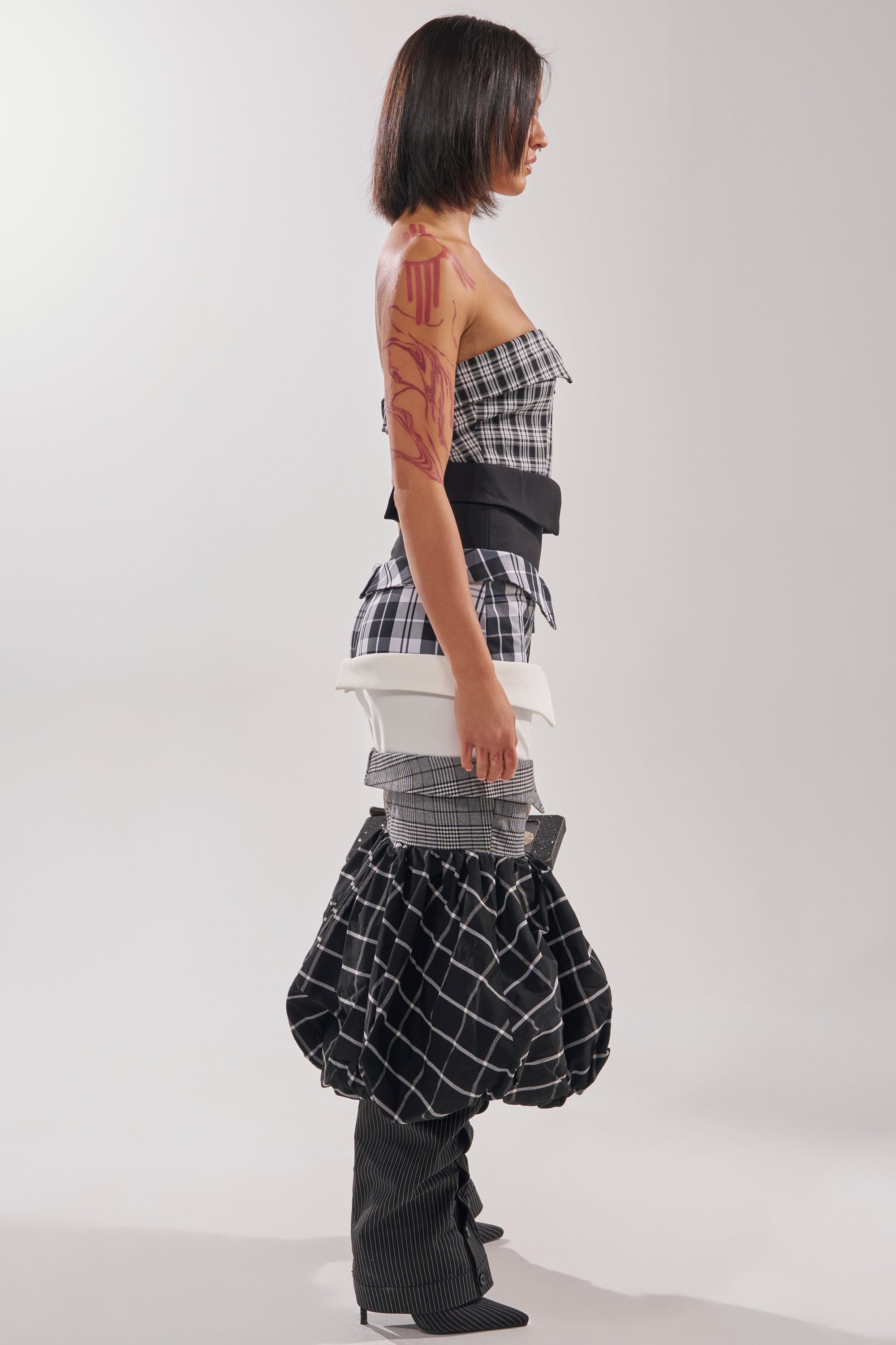 A woman in profile poses against a plain backdrop, wearing the CHANGING IT UP PLAID MIDI DRESS styled with voluminous pants, high heels, and a strapless top. She has shoulder-length straight hair and abstract red body paint on her arm.