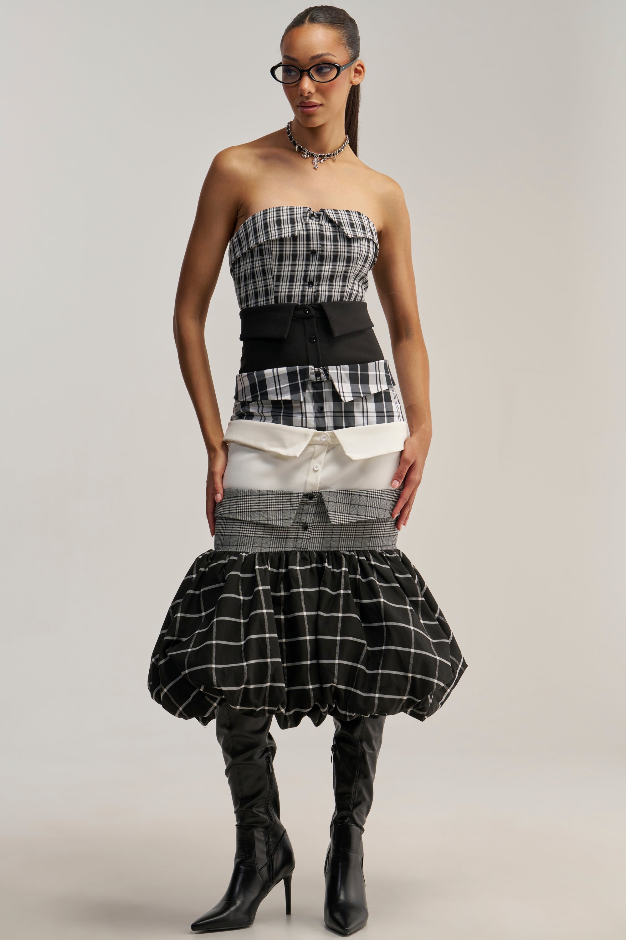 A woman wearing the CHANGING IT UP PLAID MIDI DRESS, featuring a strapless neckline and a multi-patterned, ruffled layered skirt, styled with a black belt, black heeled boots, glasses, and a necklace stands against a plain background.