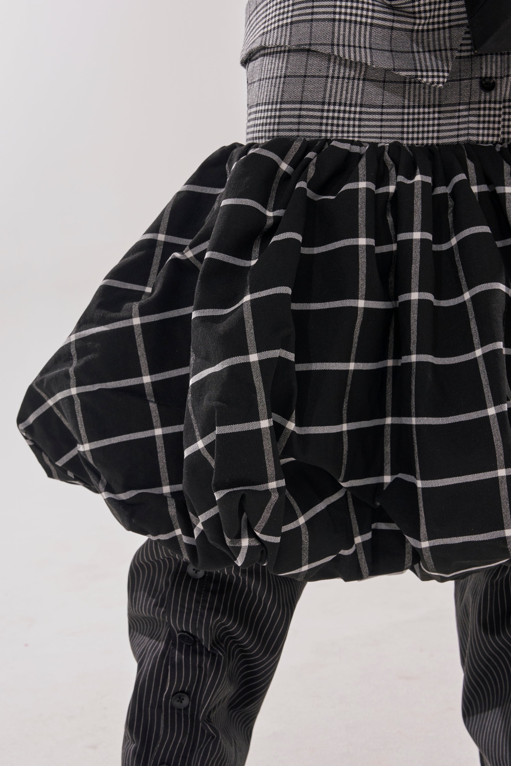 A close-up shows a person in layered outfits, featuring the CHANGING IT UP PLAID MIDI DRESS, a bold black-and-white checkered skirt, and pinstriped pants. The background is plain and light.