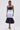 A woman wears the LOST IN THE CITY LAYERED POPLIN MIDI DRESS, designed with dark and light blue striped panels, a white waistband, and a voluminous dark skirt. She completes the look with a small white bag and white heeled sandals.