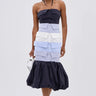 A woman wears the LOST IN THE CITY LAYERED POPLIN MIDI DRESS, designed with dark and light blue striped panels, a white waistband, and a voluminous dark skirt. She completes the look with a small white bag and white heeled sandals.