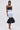 A woman stands against a plain background wearing the LOST IN THE CITY LAYERED POPLIN MIDI DRESS, which features avant-garde construction with button details and a voluminous black ruffled hem. She carries a small white handbag and wears white heeled sandals.