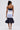 A woman with a high ponytail stands facing away, wearing the LOST IN THE CITY LAYERED POPLIN MIDI DRESS with blue, white, and striped panels and a ruffled navy hem. She carries a small white handbag and wears white heeled sandals.