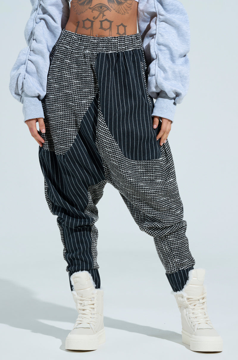 A person wearing the DO WHATEVER JOGGER in a patterned black and white style, featuring a relaxed silhouette, pairs it with a gray cropped jacket and white high-top sneakers. Their midriff reveals a tattoo as their hands rest in their pockets against the light blue background.