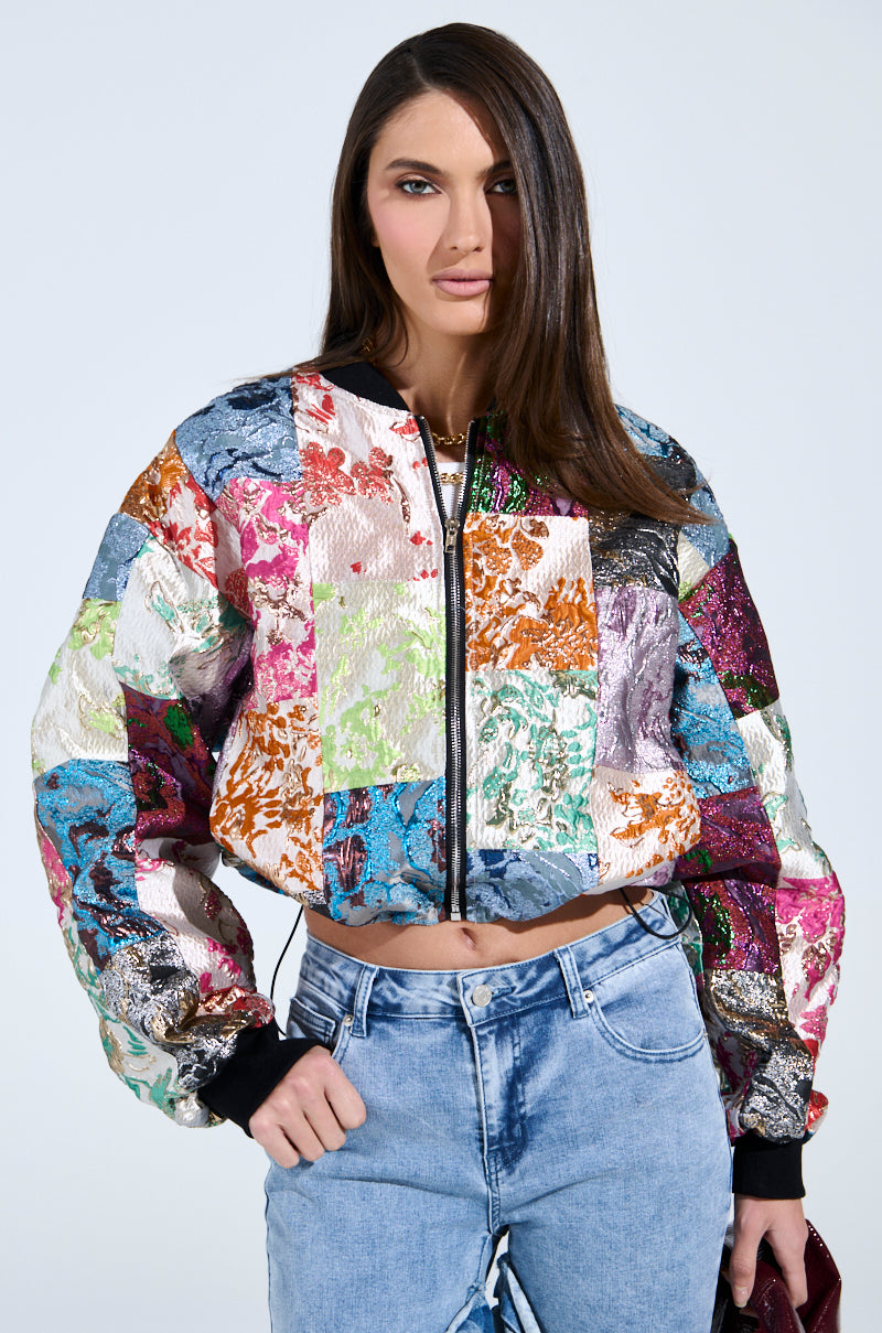 A person with long dark hair wears the THAT GIRL PATCHWORK BROCADE BOMBER, which has vibrant floral patterns, paired with light blue jeans against a plain gray background. They hold a small shiny bag.