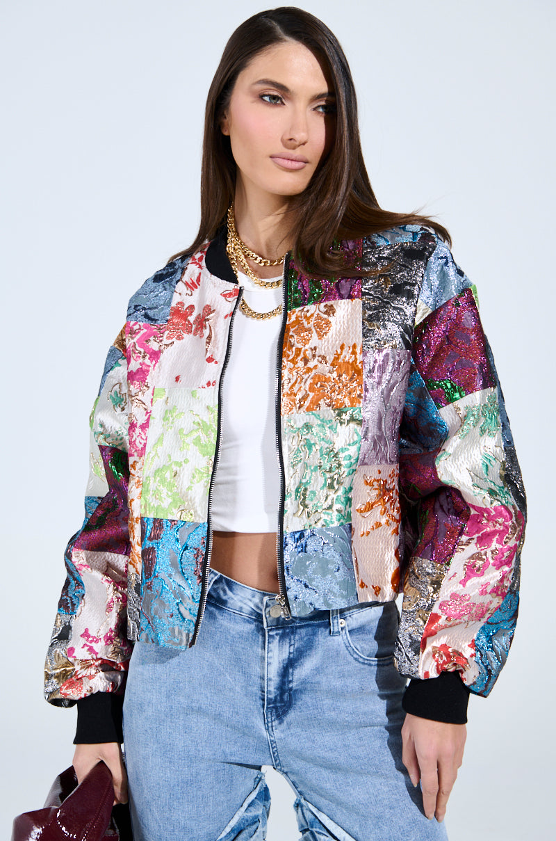 Wearing the THAT GIRL PATCHWORK BROCADE BOMBER over a white crop top and blue jeans, she carries a red purse. Her long hair highlights layered gold necklaces against a plain backdrop, crafting an effortlessly chic look.