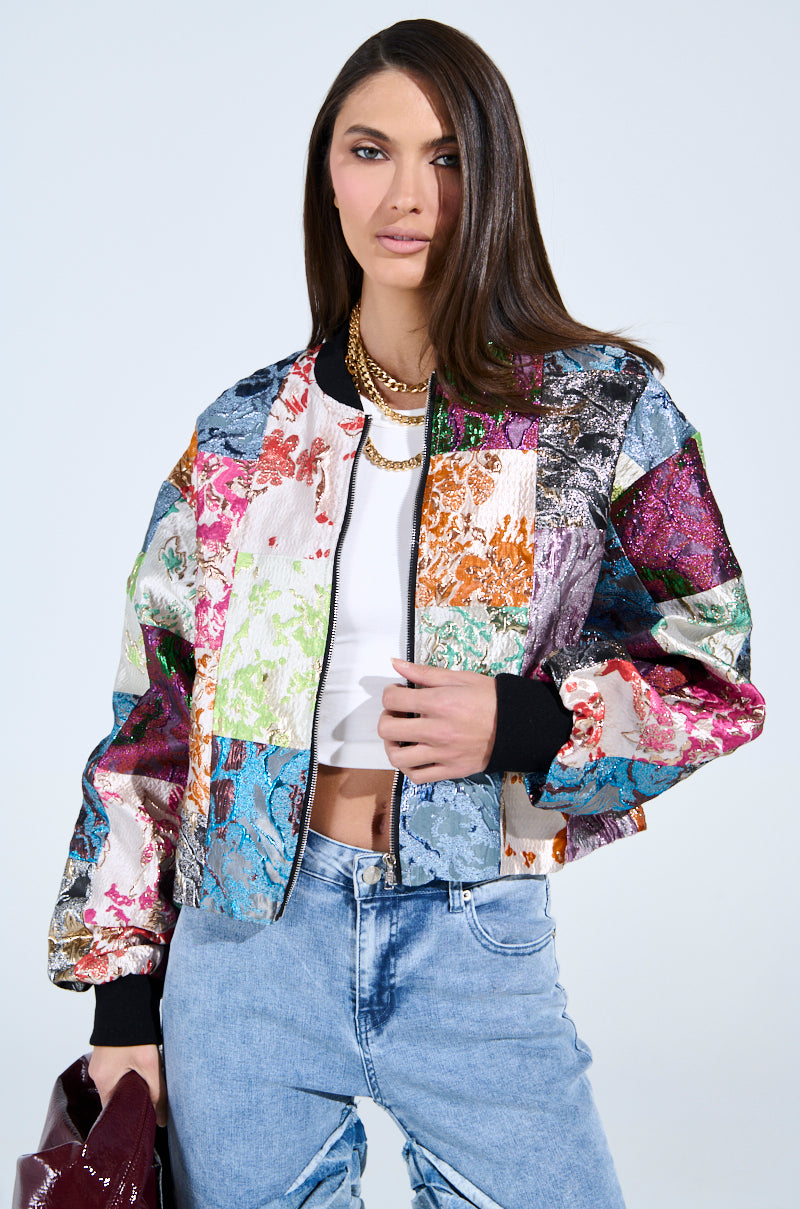 A woman with long hair wears the THAT GIRL PATCHWORK BROCADE BOMBER over a white crop top and light blue jeans. She carries a shiny maroon handbag, sports a gold chain necklace, and stands against a plain gray backdrop.