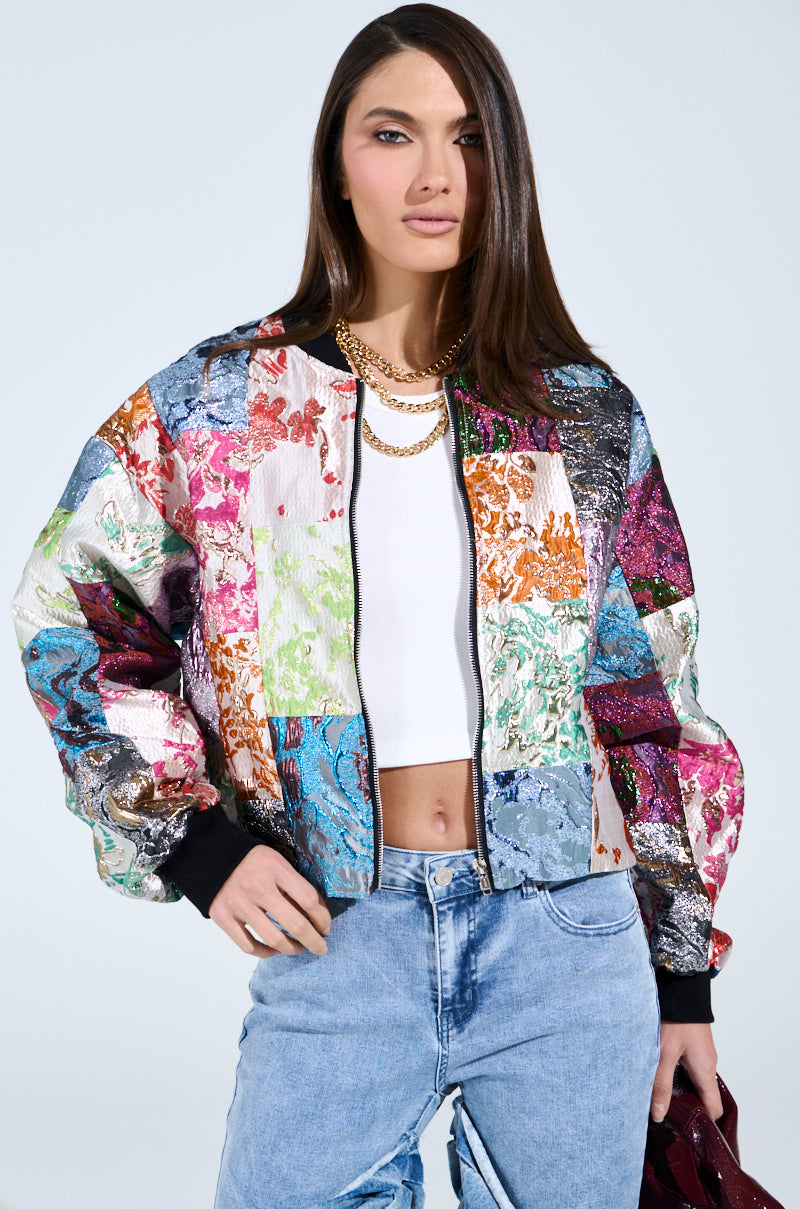 A person with long hair wears the THAT GIRL PATCHWORK BROCADE BOMBER over a white crop top and light blue jeans. Gold necklaces elegantly layer around their neck as they hold a red object, with a simple, light backdrop highlighting their vibrant attire.