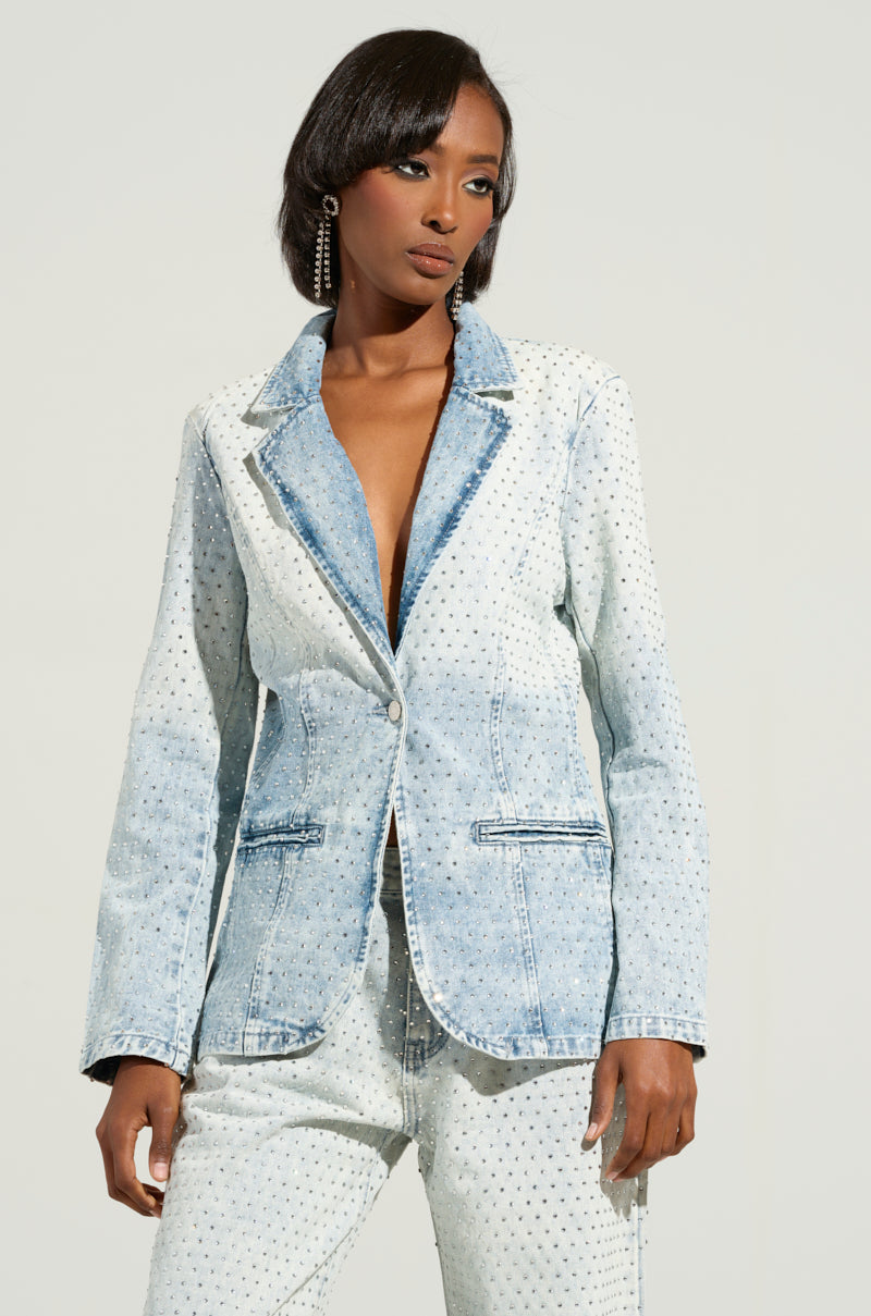 Wearing the ELECTRIC EMBELLISHED DENIM BLAZER in a light blue wash, a person poses against a plain background. The chic ensemble features matching jeans and delicate rhinestone embellishments. Styled with short hair and hoop earrings, they exude an effortlessly fashionable vibe.