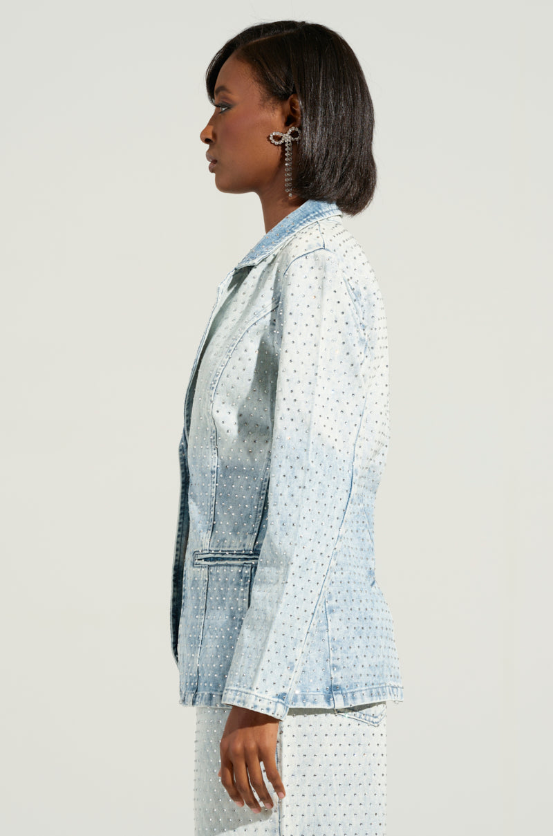 A person with short dark hair is shown in profile, wearing the ELECTRIC EMBELLISHED DENIM BLAZER, a light blue blazer adorned with rhinestone embellishments. They sport an intricate earring, and the soft gray background enhances the chic business casual vibe.