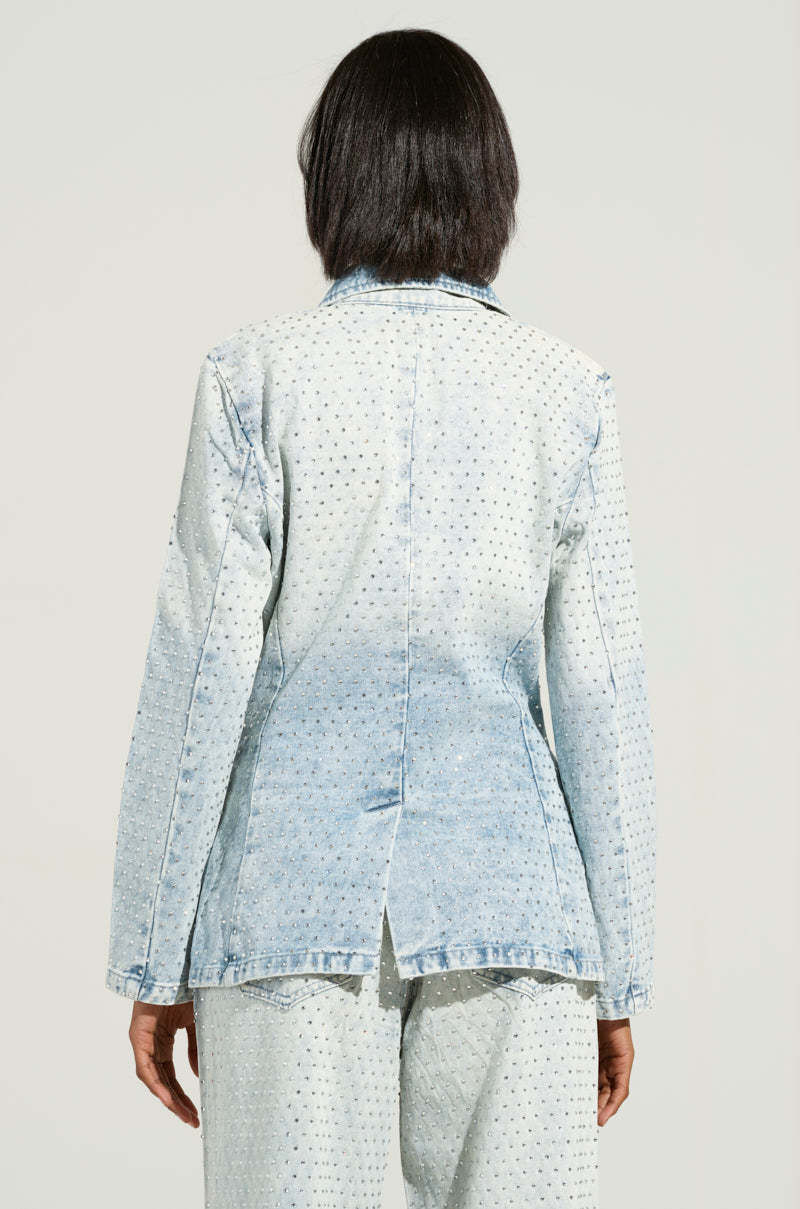 A rear view showcases an individual donning the ELECTRIC EMBELLISHED DENIM BLAZER, a light blue denim jacket adorned with rhinestones. Styled like a casual blazer, it is paired with matching pants and features a split hem at the back, set against a plain, light-colored background.