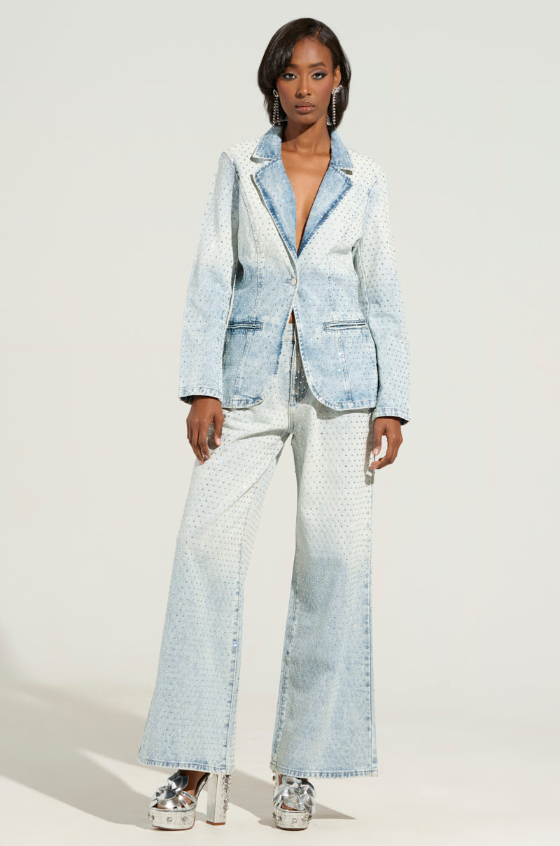 Against a plain background, a woman radiates confidence in her business casual ensemble. She dons an ELECTRIC EMBELLISHED DENIM BLAZER with flared pants and matching platform shoes. Her impeccably styled hair further elevates her poised and polished appearance.