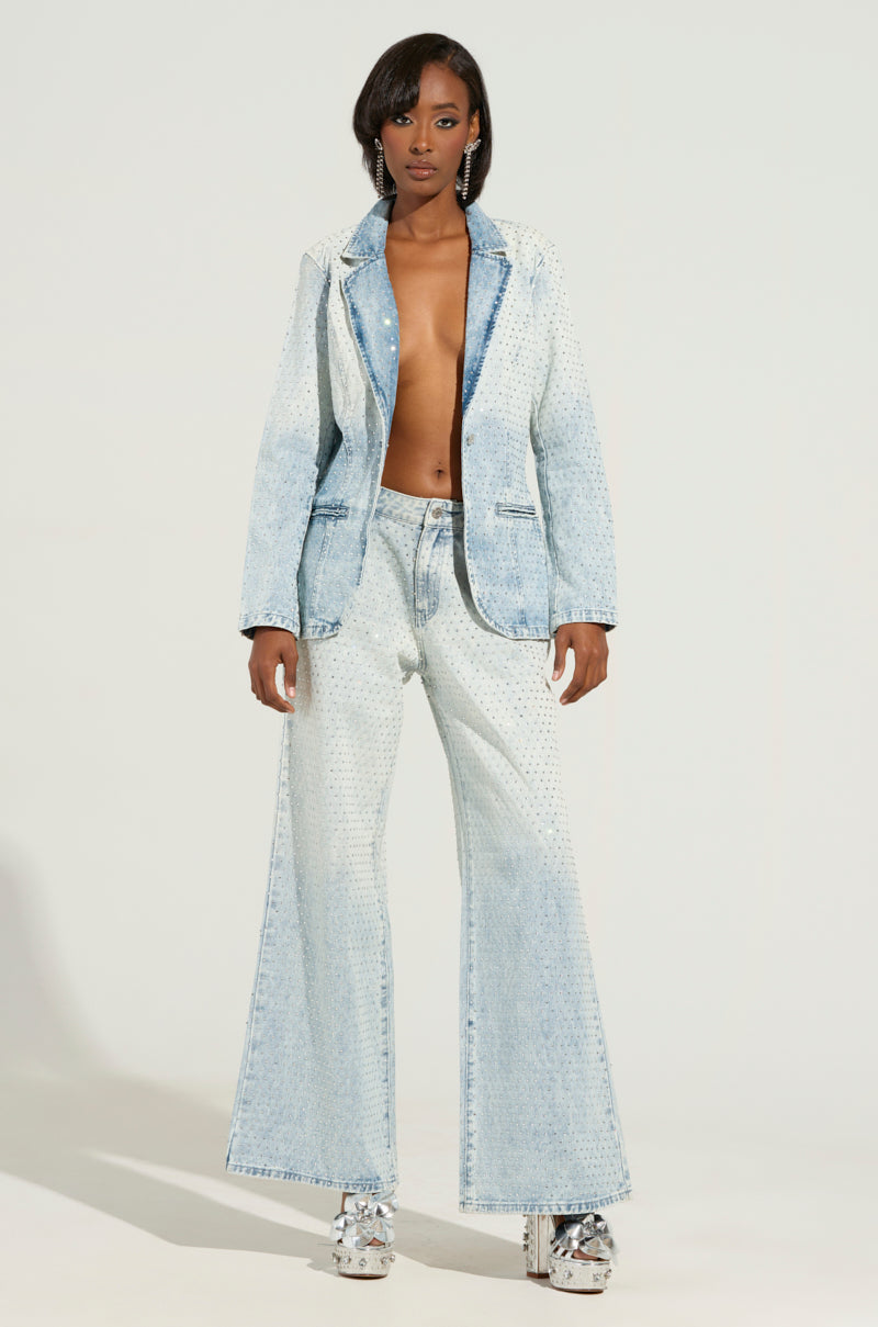 A person confidently models the ELECTRIC EMBELLISHED DENIM BLAZER, a stylish, light blue piece paired with flared pants. Rhinestone embellishments add flair to the outfit, while platform shoes complete the look. The blazer is worn open, revealing bare skin beneath, all set against a plain white backdrop that blends business casual elegance with daring style.