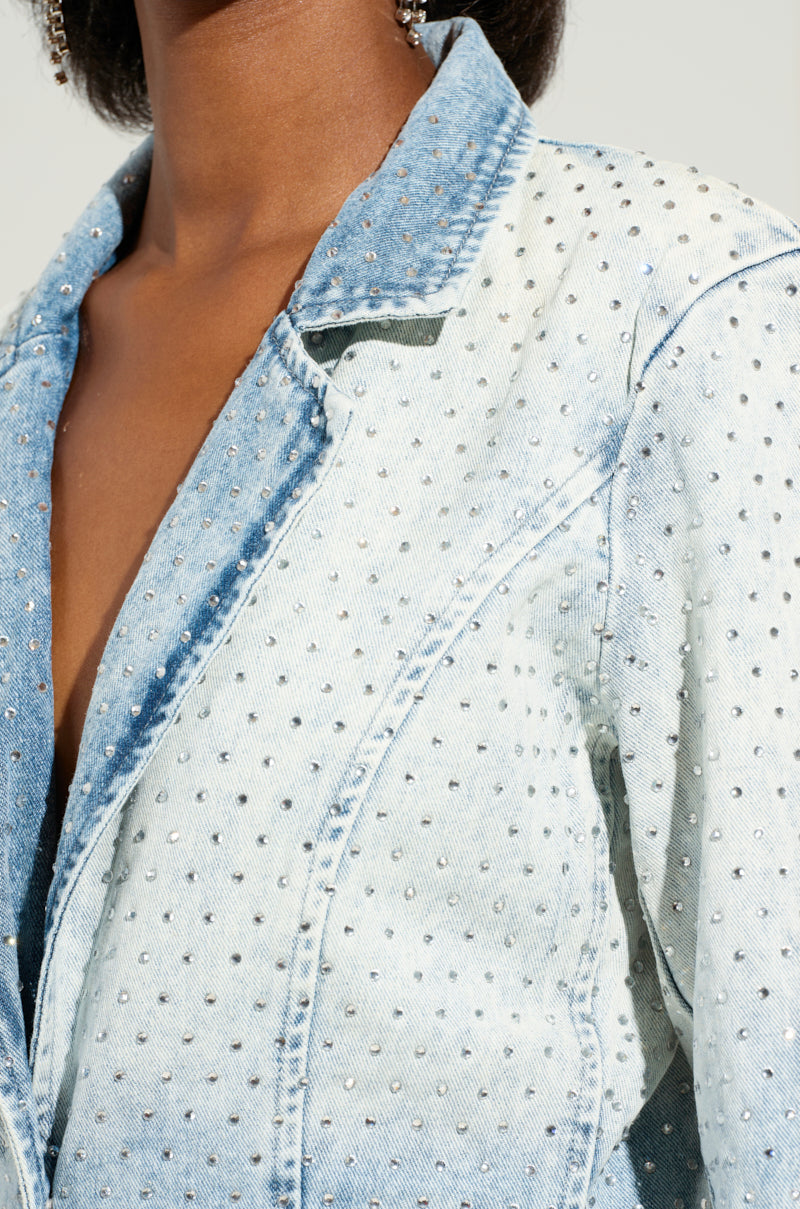 A close-up of the ELECTRIC EMBELLISHED DENIM BLAZER reveals a textured pattern created by numerous small studs. The blazer features a light blue wash, making it ideal for a business casual outfit. The individual's upper body is partially visible, but their face remains out of view.