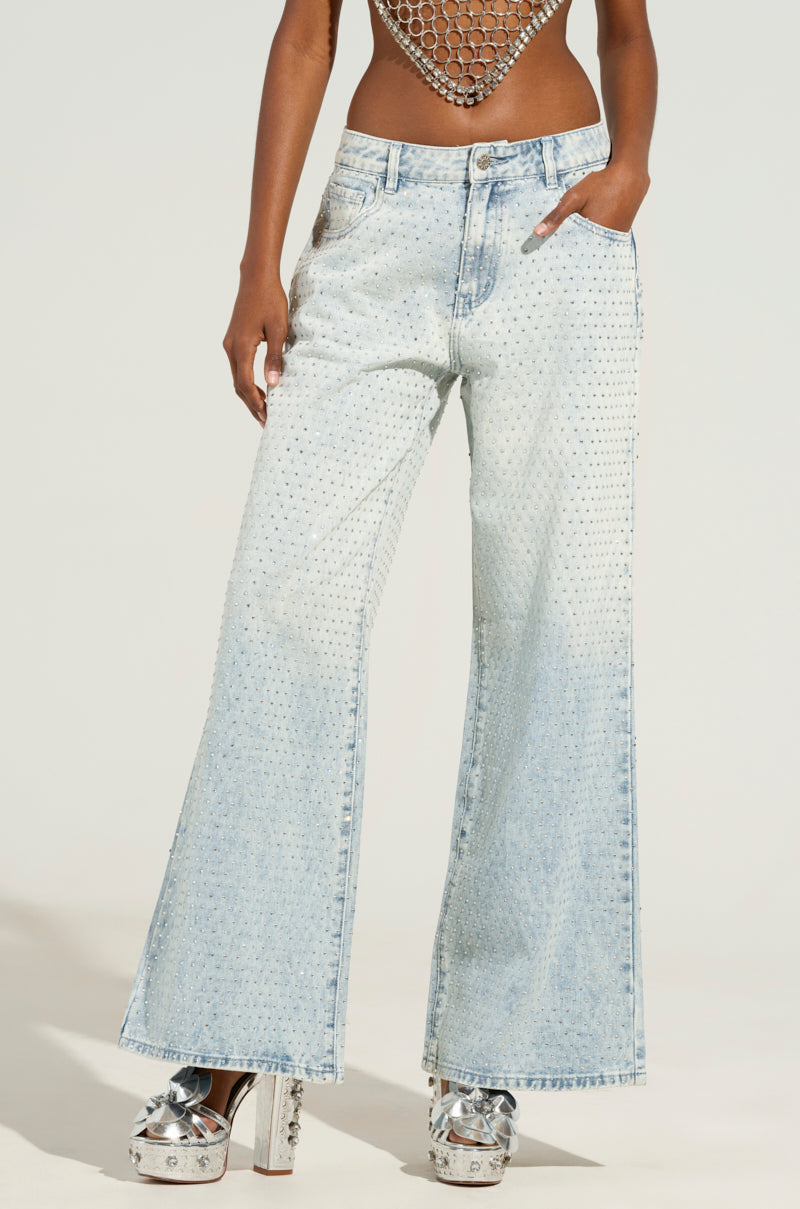Wearing the ELECTRIC EMBELLISHED WIDE LEG JEANS in a light wash denim style, adorned with rhinestones throughout, they effortlessly pair them with a mesh top and silver platform shoes against a plain backdrop that perfectly highlights their striking ensemble.