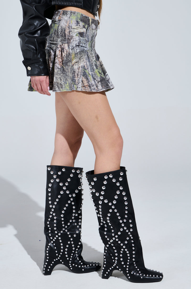 Someone wearing the MOVIN DIFFERENT MINI SKIRT, a camo-patterned mini with a high-rise fit, pairs it with a black leather jacket and knee-high boots adorned with silver studs. The subtle gray backdrop complements the look perfectly.