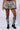 A person wearing a MARIAH TREE CAMO CARGO MINI SKIRT with camo pattern and cargo pockets stands with their hands resting on the skirt. They are also sporting tall, houndstooth-patterned socks paired with black chunky boots, against a plain white background.