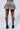 A person stands facing away, showcasing the back of the MARIAH TREE CAMO CARGO MINI SKIRT, a pleated design in shades of grey and yellow. They are paired with black studded knee-high boots featuring straps and buckles. The plain white background highlights the outfit's bold statement, reminiscent of the edgy designs from AKIRA Label.