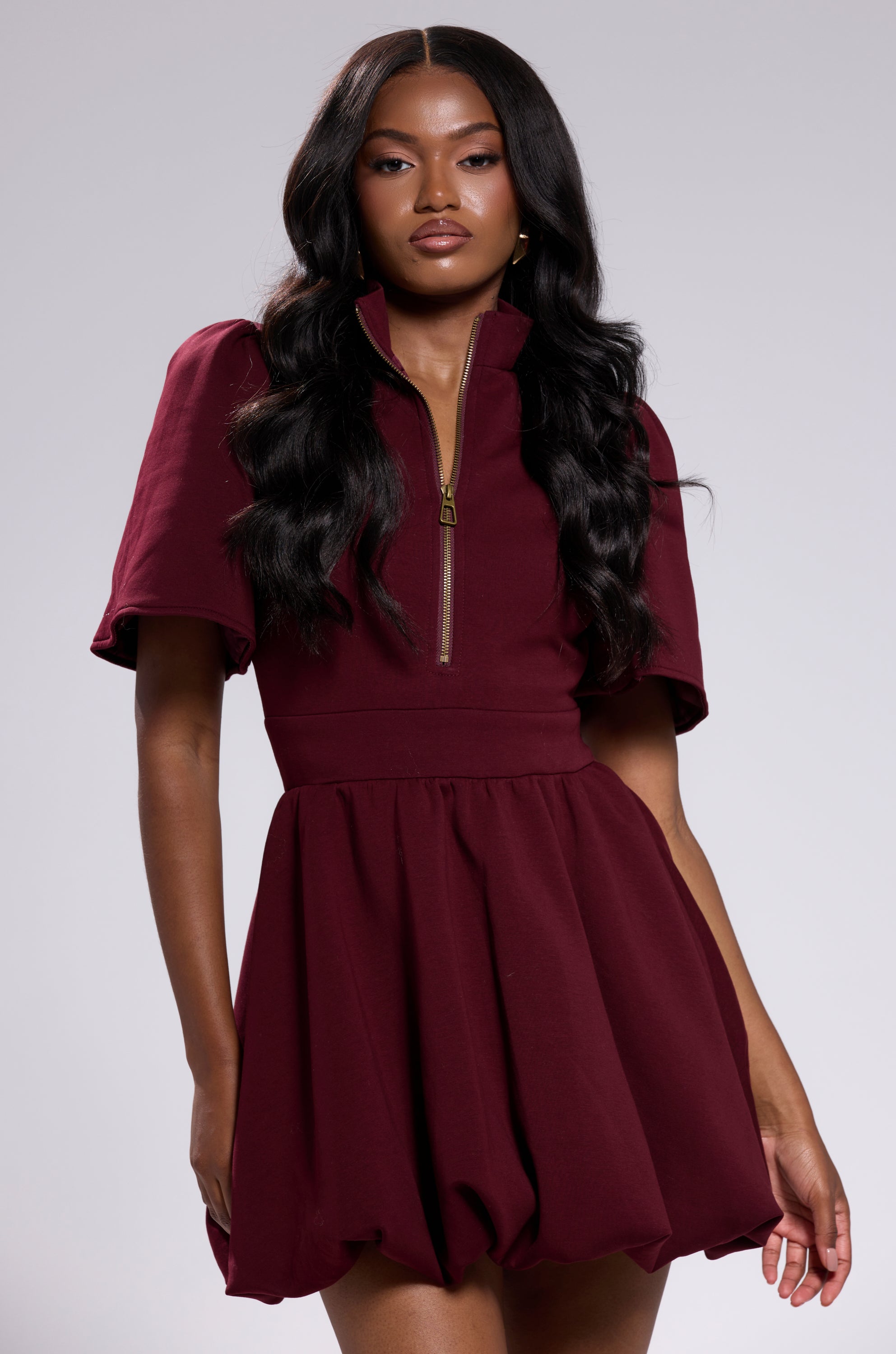 A woman with long, wavy black hair models the NAME OF THE GAME SHORT SLEEVE ZIPPERED MINI DRESS IN BURGUNDY, featuring a flared skirt, in front of a plain light gray background.