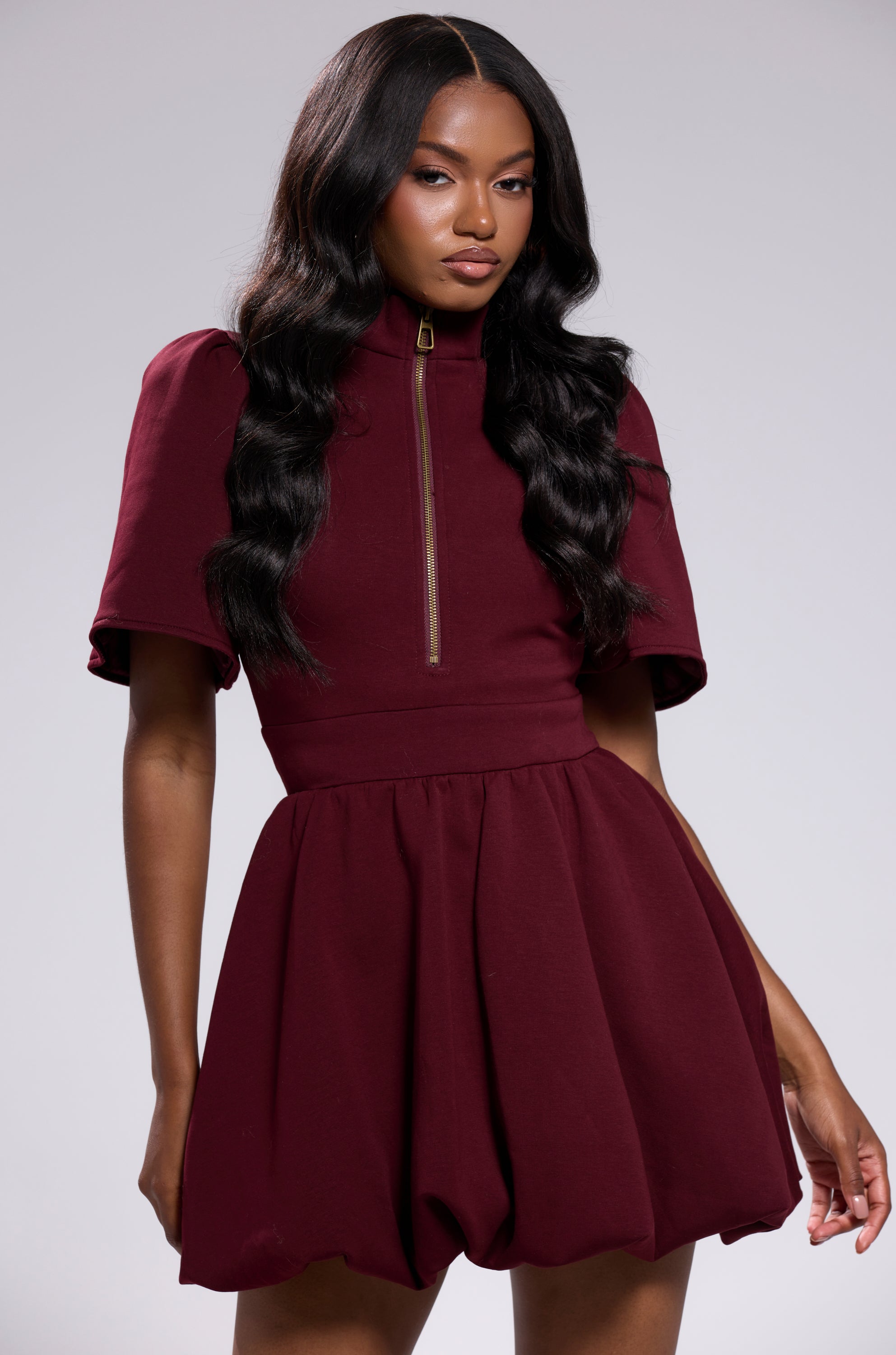 A woman with long, wavy black hair models the NAME OF THE GAME SHORT SLEEVE ZIPPERED MINI DRESS IN BURGUNDY, featuring a zip-up high collar and flared skirt, against a light gray background.