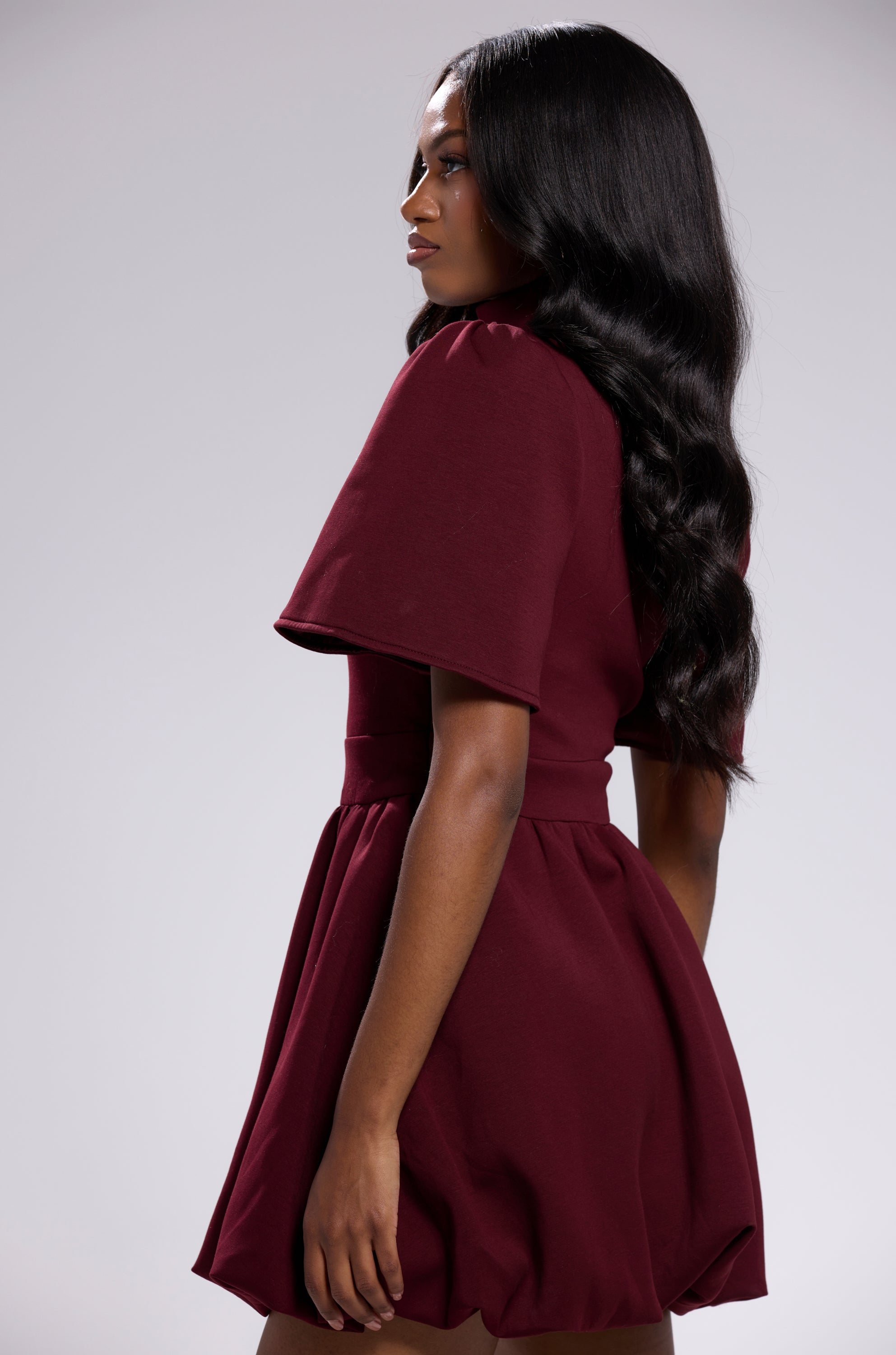 A woman with long, wavy hair stands sideways against a plain light gray background, wearing the NAME OF THE GAME SHORT SLEEVE ZIPPERED MINI DRESS IN BURGUNDY featuring a flared skirt and short sleeves.