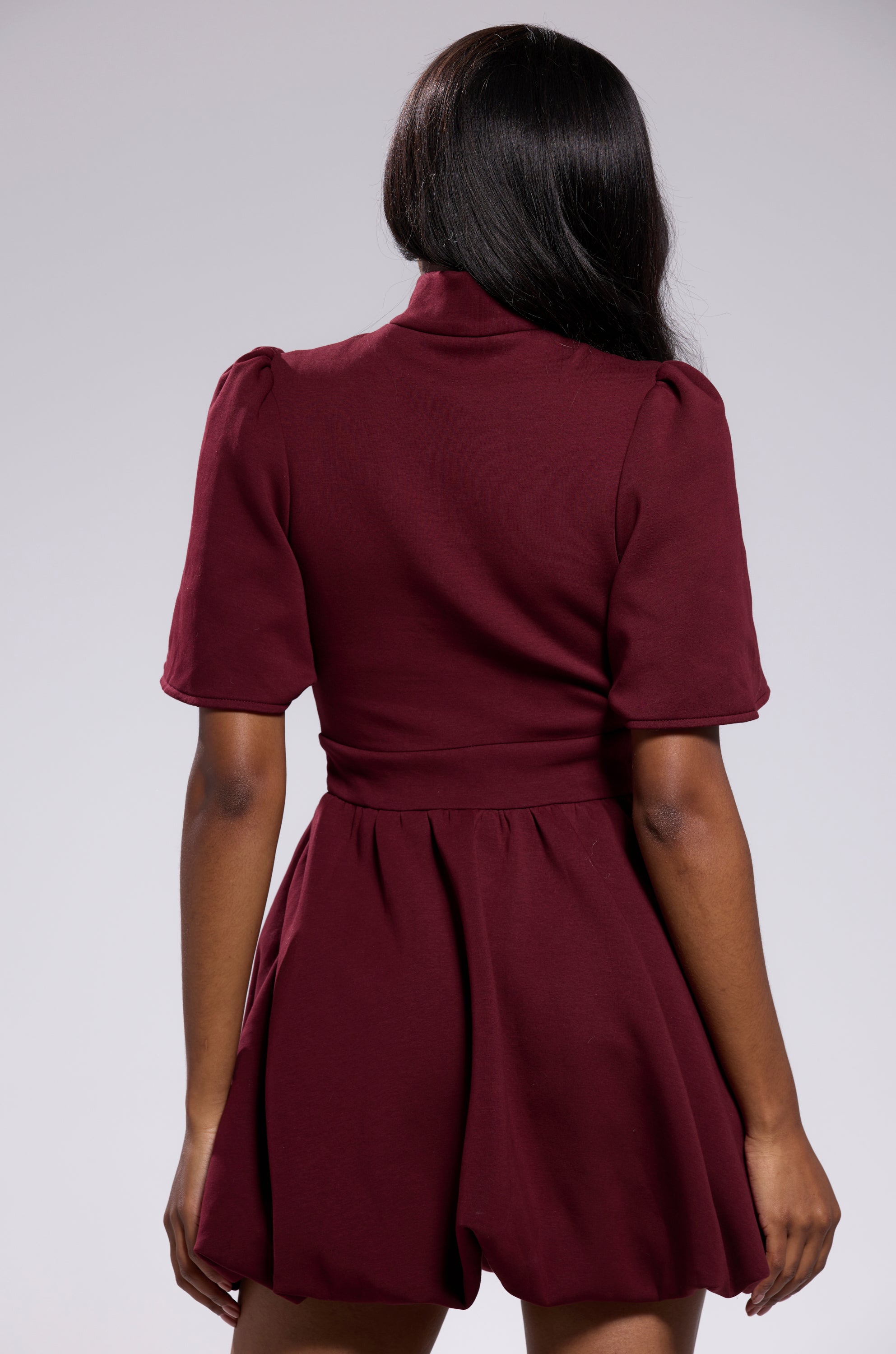 A woman with long dark hair stands with her back to the camera, wearing the NAME OF THE GAME SHORT SLEEVE ZIPPERED MINI DRESS IN BURGUNDY, featuring a fitted waist and flared skirt, against a plain light background.