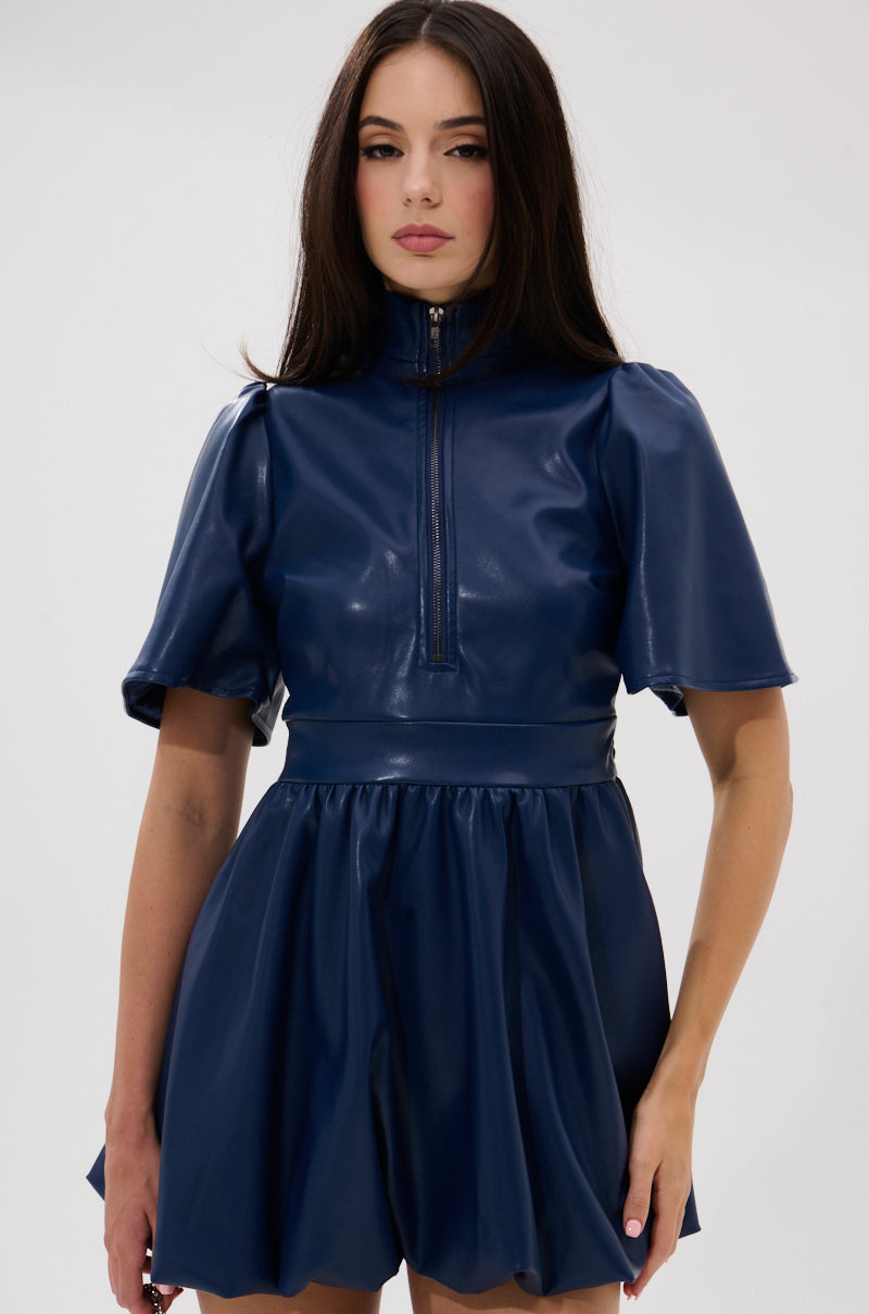 A woman with long dark hair models the LALA LOVE YOU PU MINI DRESS IN NAVY, featuring short sleeves, a high neck, a zippered front, and a flared skirt, standing against a plain white background.