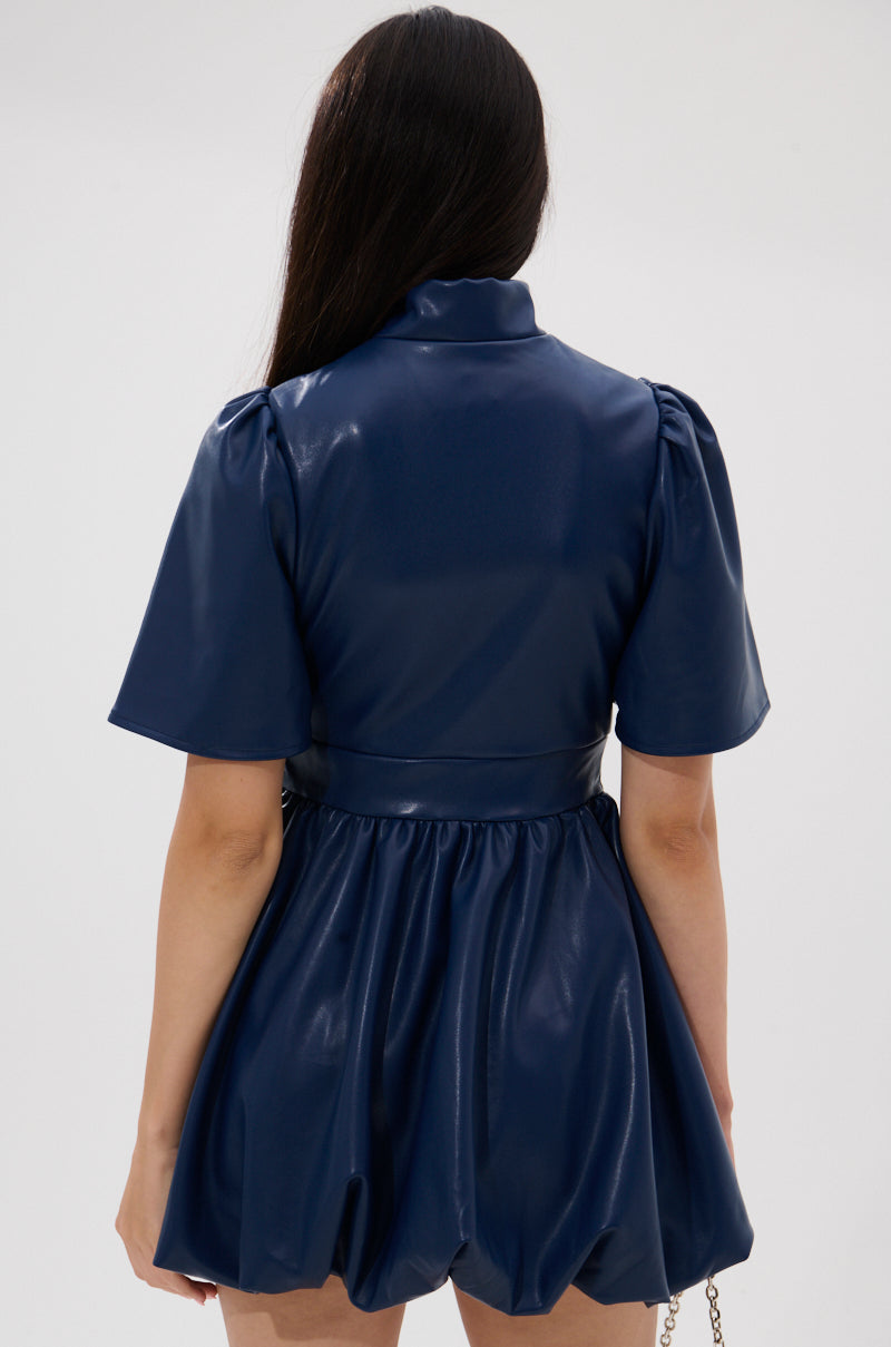A woman with long dark hair stands with her back to the camera, wearing the LALA LOVE YOU PU MINI DRESS IN NAVY—a short-sleeved faux leather dress with a flared, gathered skirt—against a plain light background.
