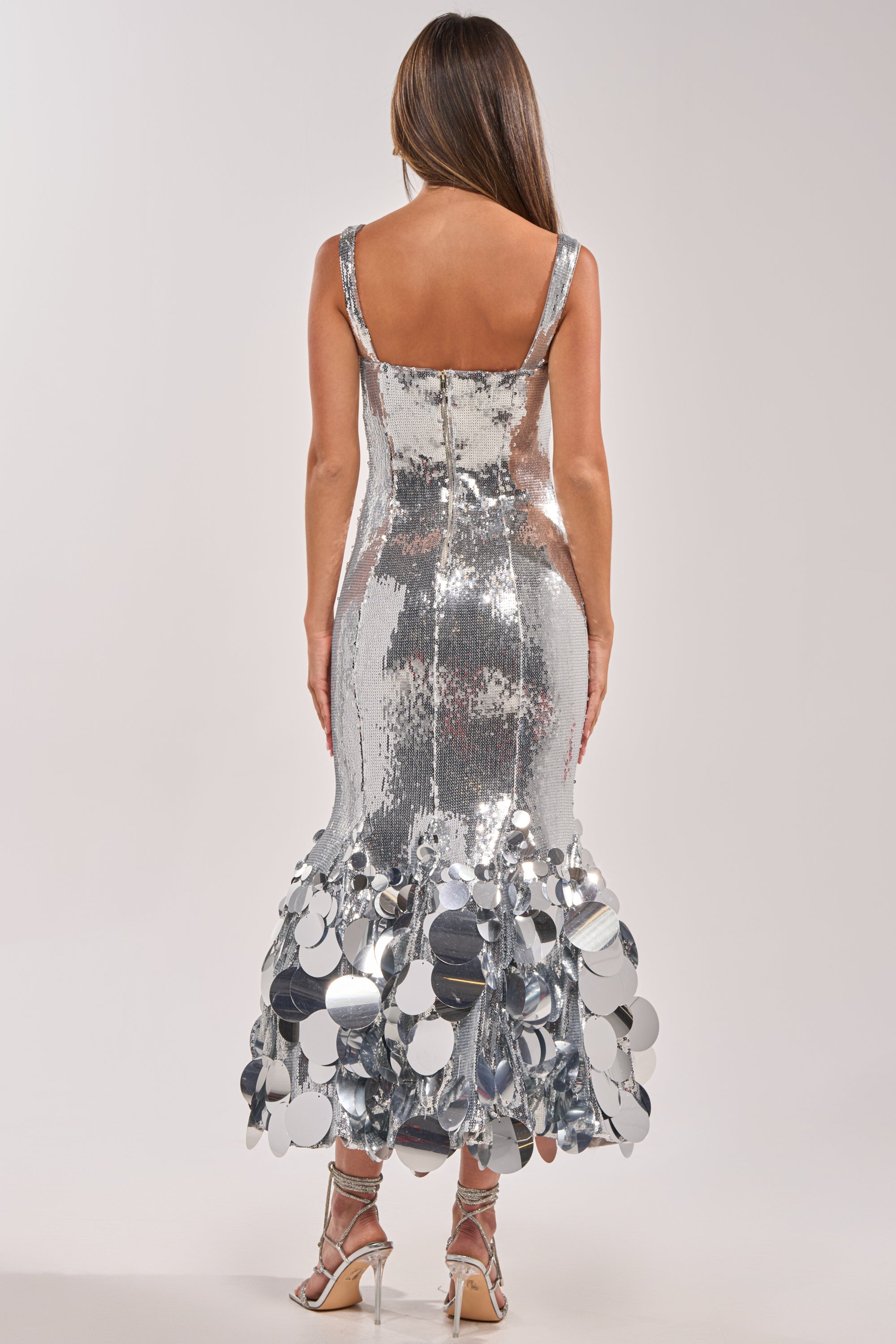 A woman faces away in the LIFE OF THE PARTY SEQUIN MIDI DRESS IN SILVER, featuring a sequin fishtail hem with large metallic discs. Paired with strappy heels, her long hair flows down against a plain background—a bold formal look. 3