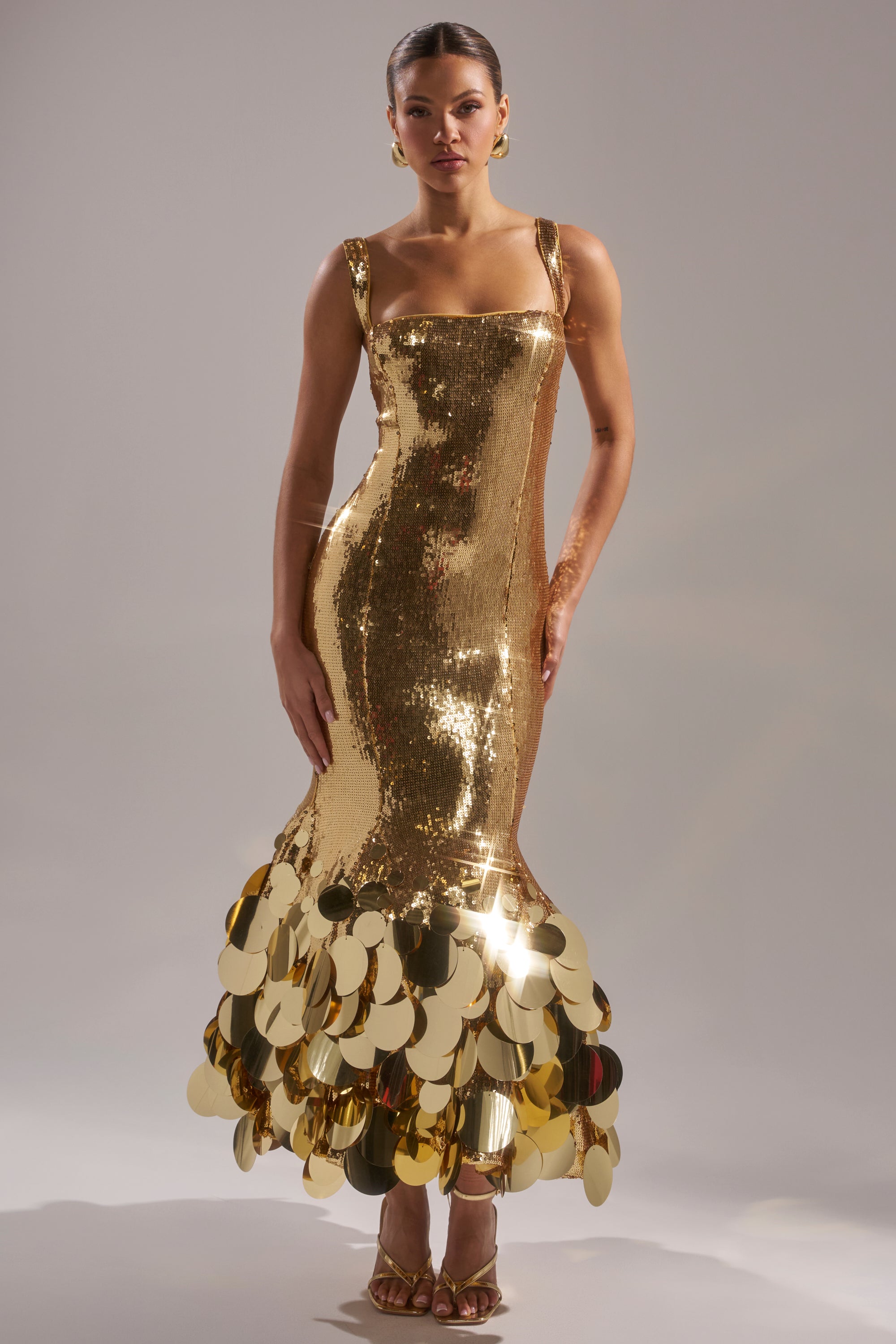 A woman stands against a light background wearing the LIFE OF THE PARTY SEQUIN MIDI DRESS, a fitted, sleeveless gold sequin dress with bold gold discs at the hem. Her hair is pulled back, and she accessorizes with gold heels and earrings.