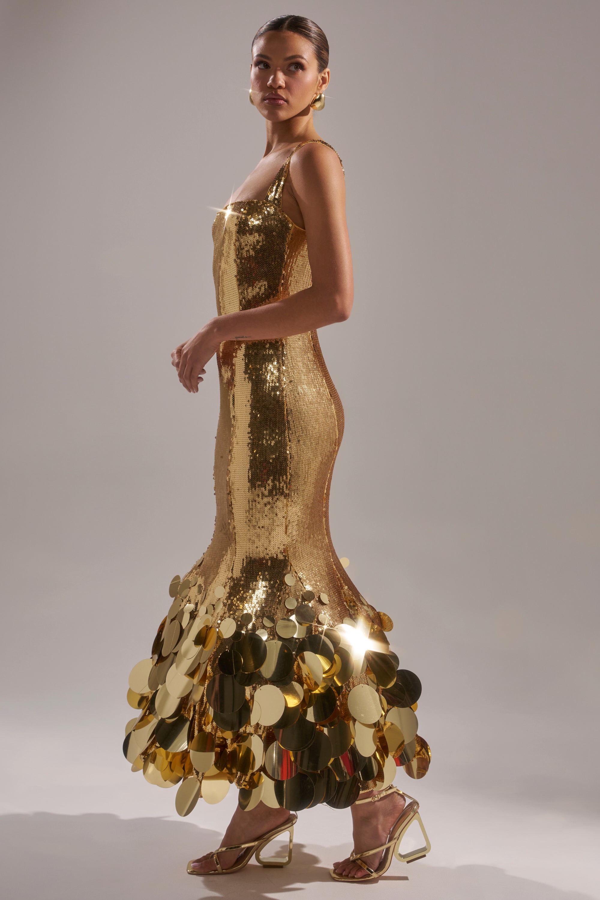 A woman poses in the LIFE OF THE PARTY SEQUIN MIDI DRESS, a fitted sleeveless gold sequin gown with large gold discs at the hem, styled with gold high-heeled sandals against a plain light background.
