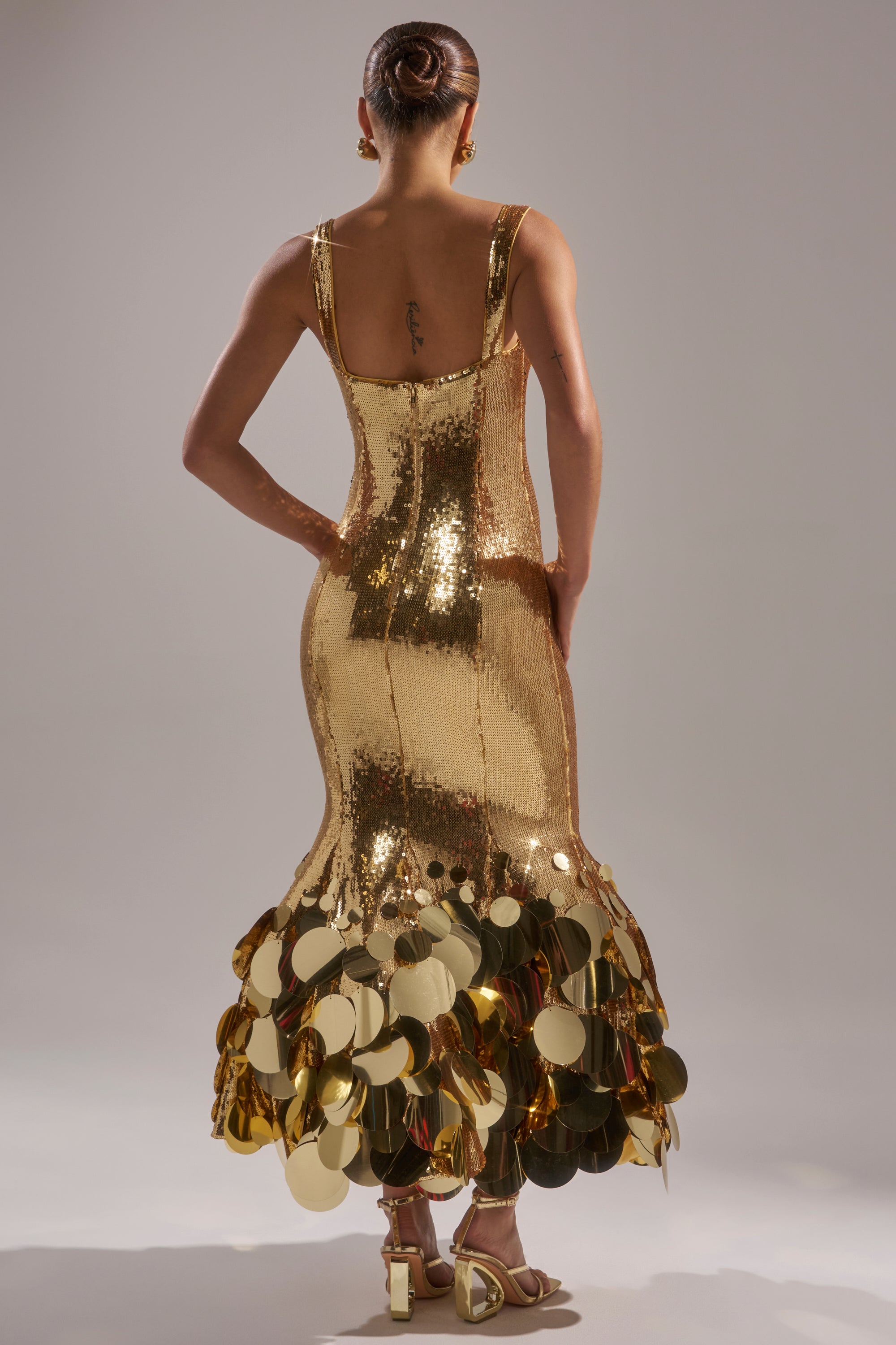 A woman stands facing away in the LIFE OF THE PARTY SEQUIN MIDI DRESS—shiny gold with large discs at the hem. Her hair is in a neat bun, she wears gold high-heeled sandals, and the softly lit background is plain.