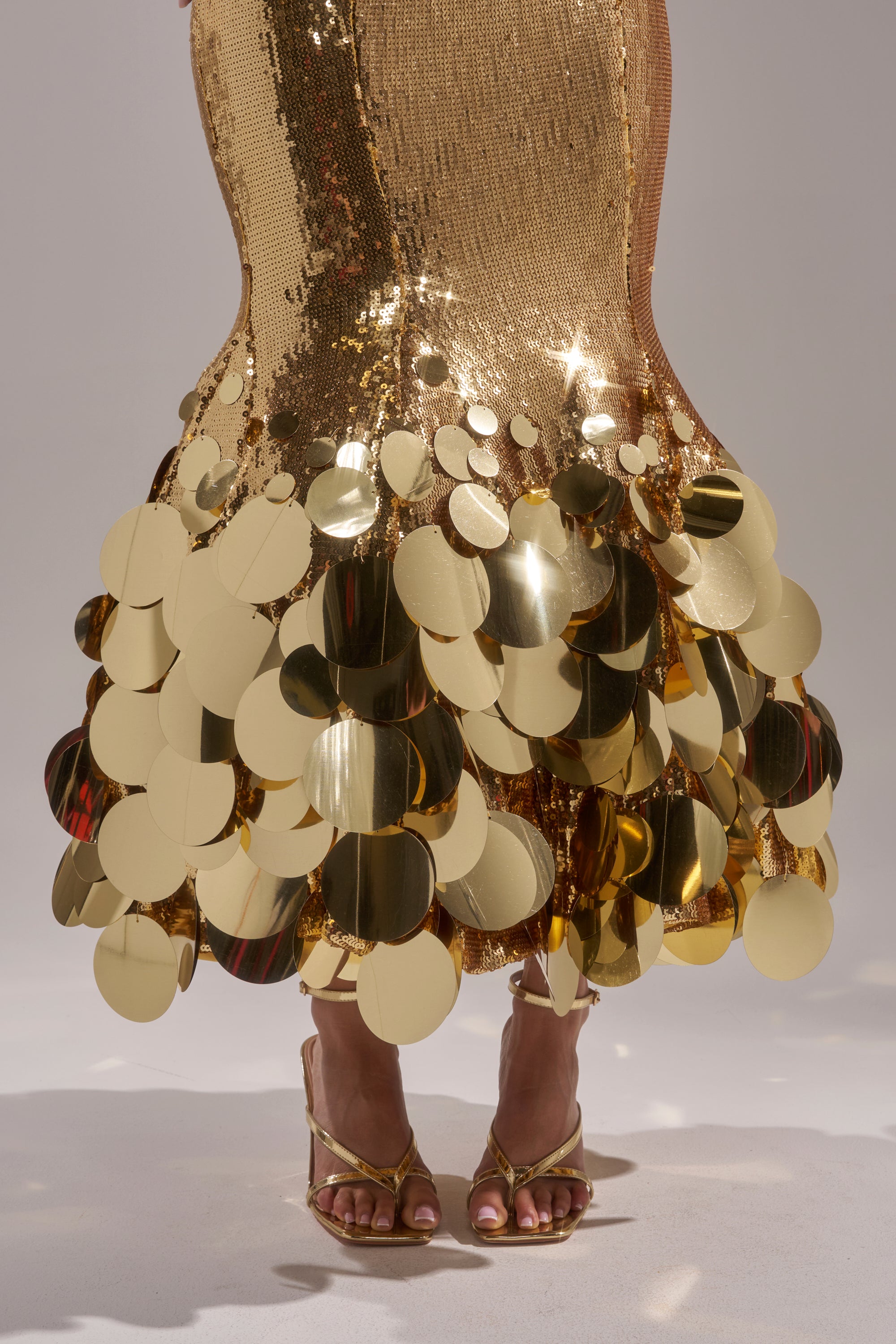 A person wearing the LIFE OF THE PARTY SEQUIN MIDI DRESS, featuring shiny gold sequins with large reflective discs at the hem, and gold high-heeled sandals. Only the lower legs and feet are visible.