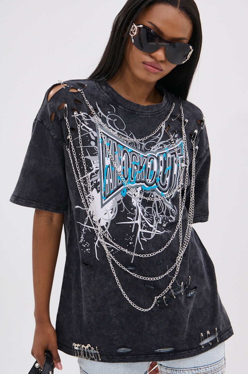 A woman in large black sunglasses and the KNOCK YOURSELF OUT DISTRESSED TEE, featuring chain and safety pin details, ripped shoulders, and a bold blue-and-white graphic. Her straight hair and confident look complete the grungy-chic vibe.