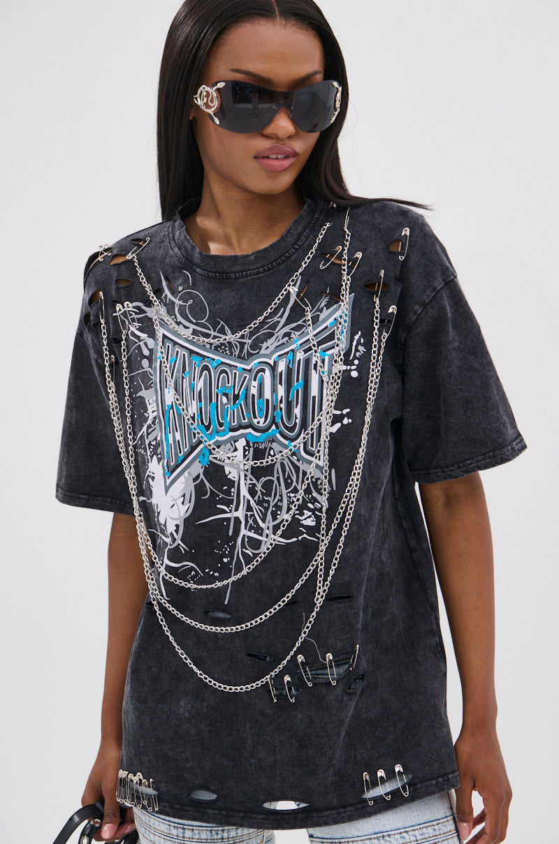 A woman sports the KNOCK YOURSELF OUT DISTRESSED TEE with silver chains and safety pins, styling it grungy-chic. She has straight hair, large sunglasses, a small purse, and stands against a plain light background.