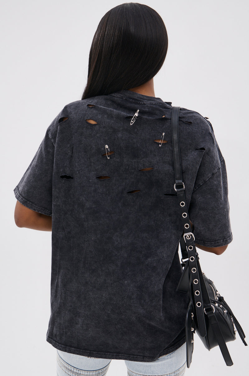 Person with long straight hair, seen from behind, wears the KNOCK YOURSELF OUT DISTRESSED TEE with rips and safety pins for a grungy-chic look. They carry a black shoulder bag with metal details and wear light blue jeans.