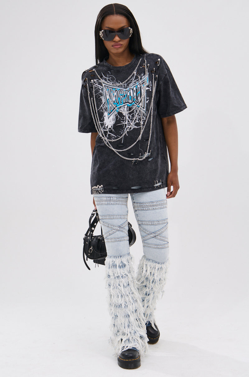 A person wearing dark sunglasses, the KNOCK YOURSELF OUT DISTRESSED TEE with chains, light blue acid-washed crisscross jeans, furry white leg warmers, and holding a small black bag walks forward against a plain background.
